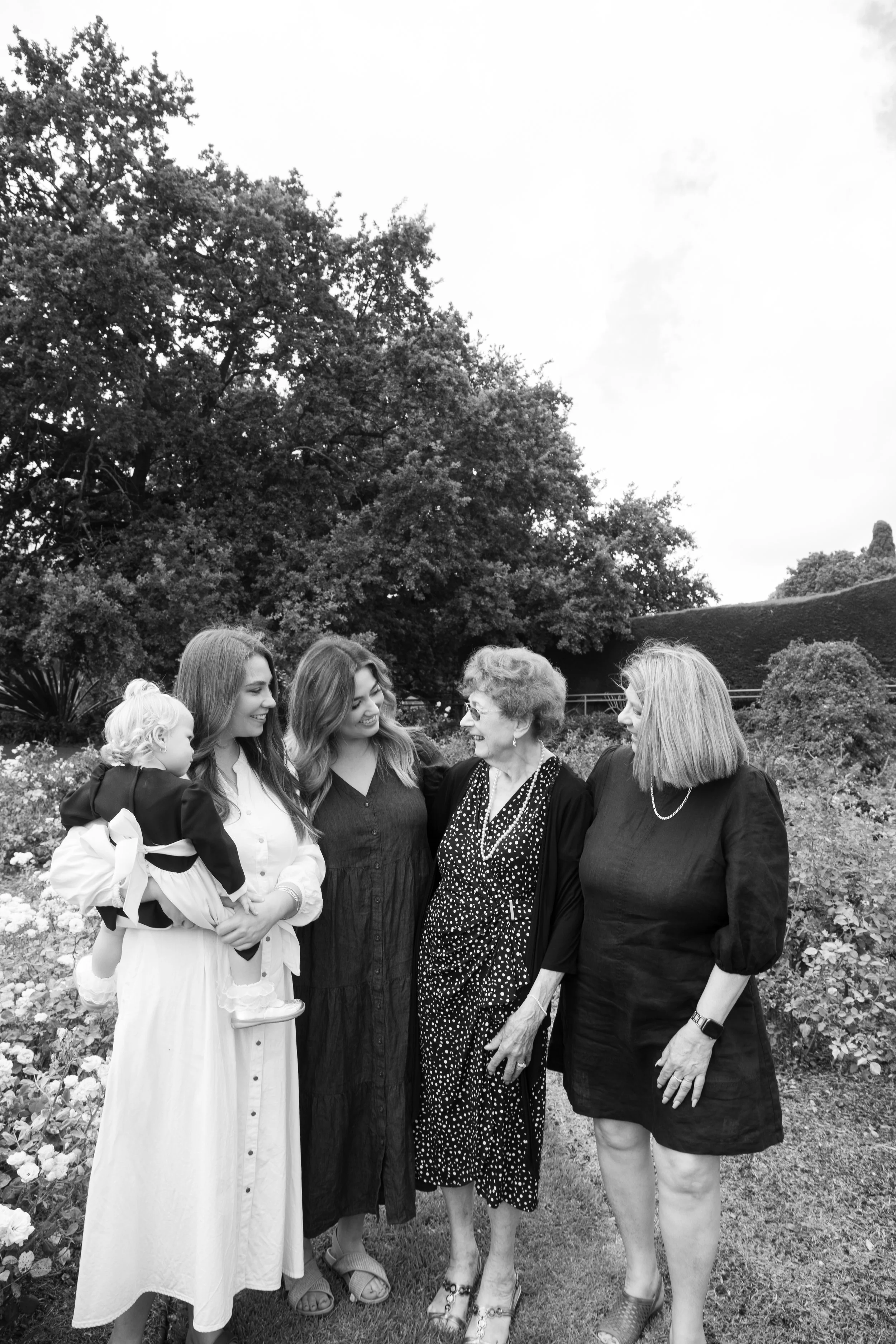 Five women, including a young girl holding a toddler, stand together outdoors in a garden, engaging in conversation and smiling.