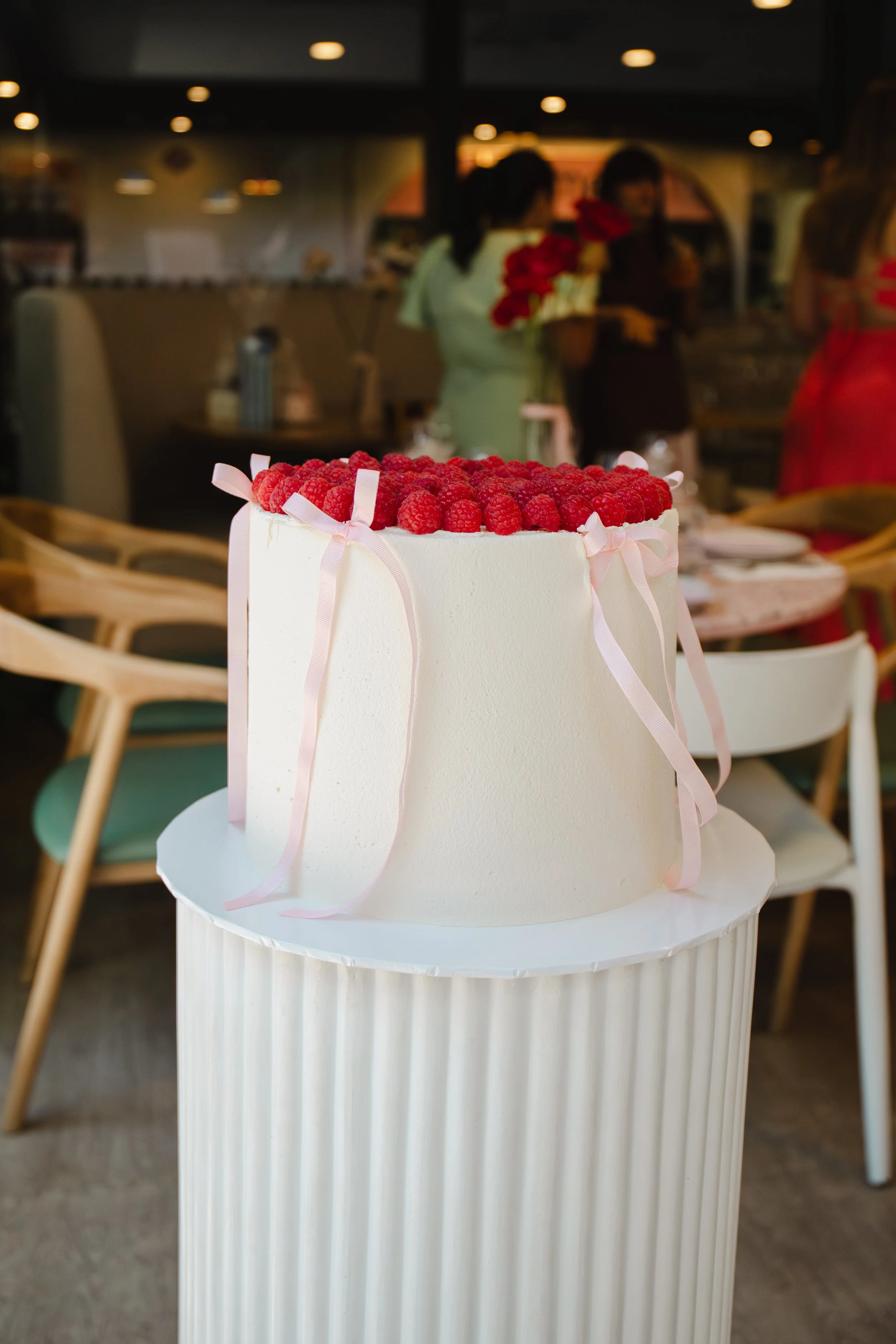 A white cake decorated with raspberries and pink ribbons on top, placed on a white pedestal in a restaurant setting with people in the background.