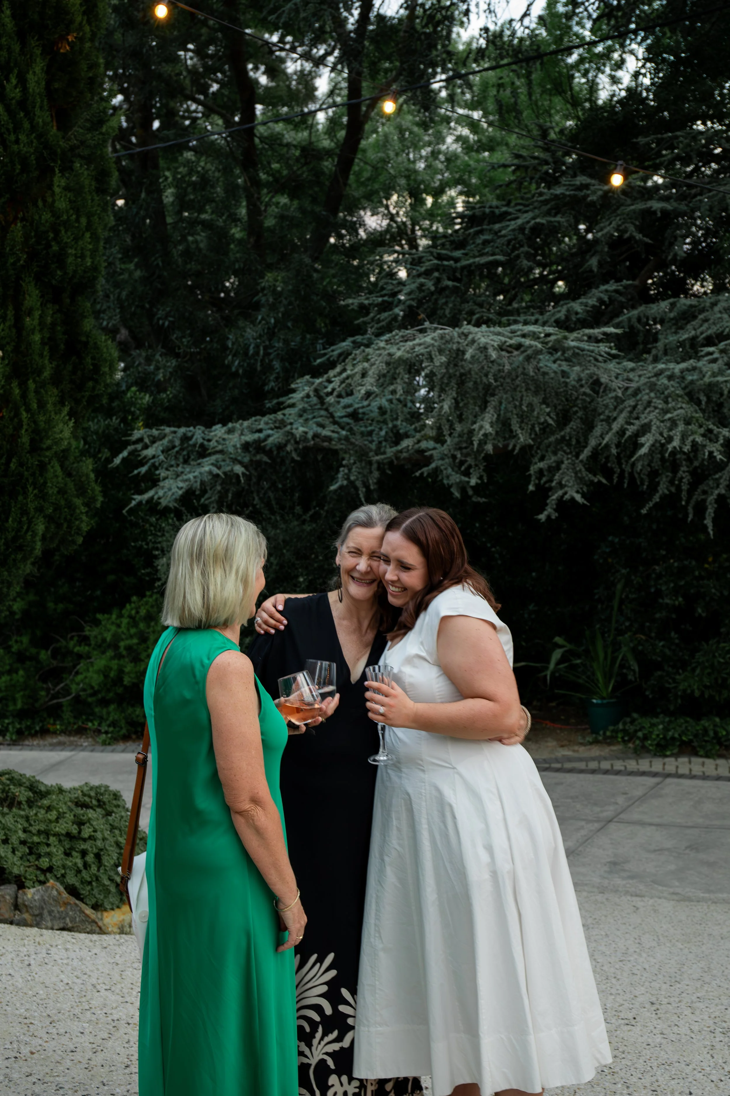 Three women socializing outdoors at dusk, holding wine glasses, surrounded by trees and string lights.