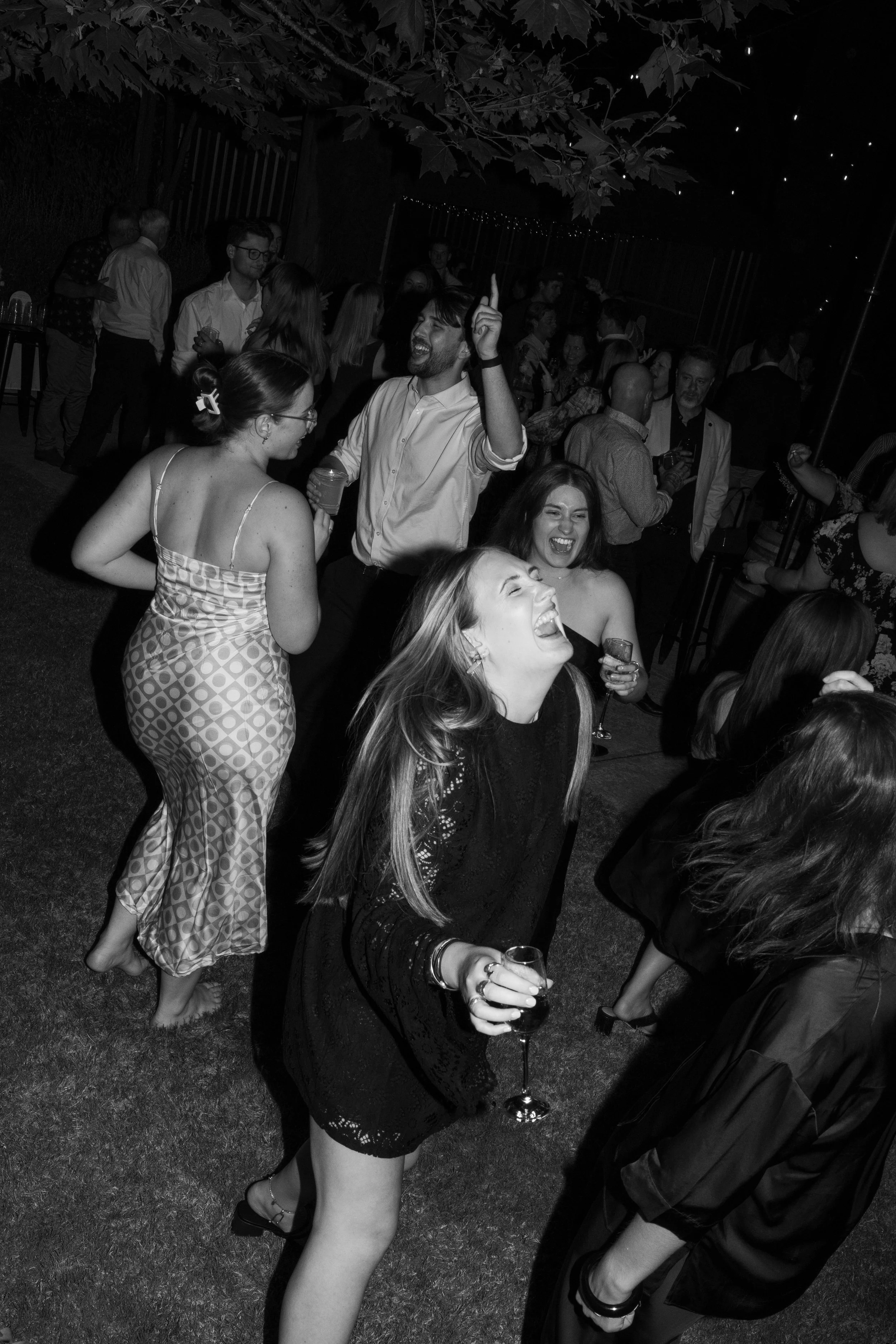 People dancing and socializing at a party or celebration outdoors at night, some holding drinks, with string lights overhead.