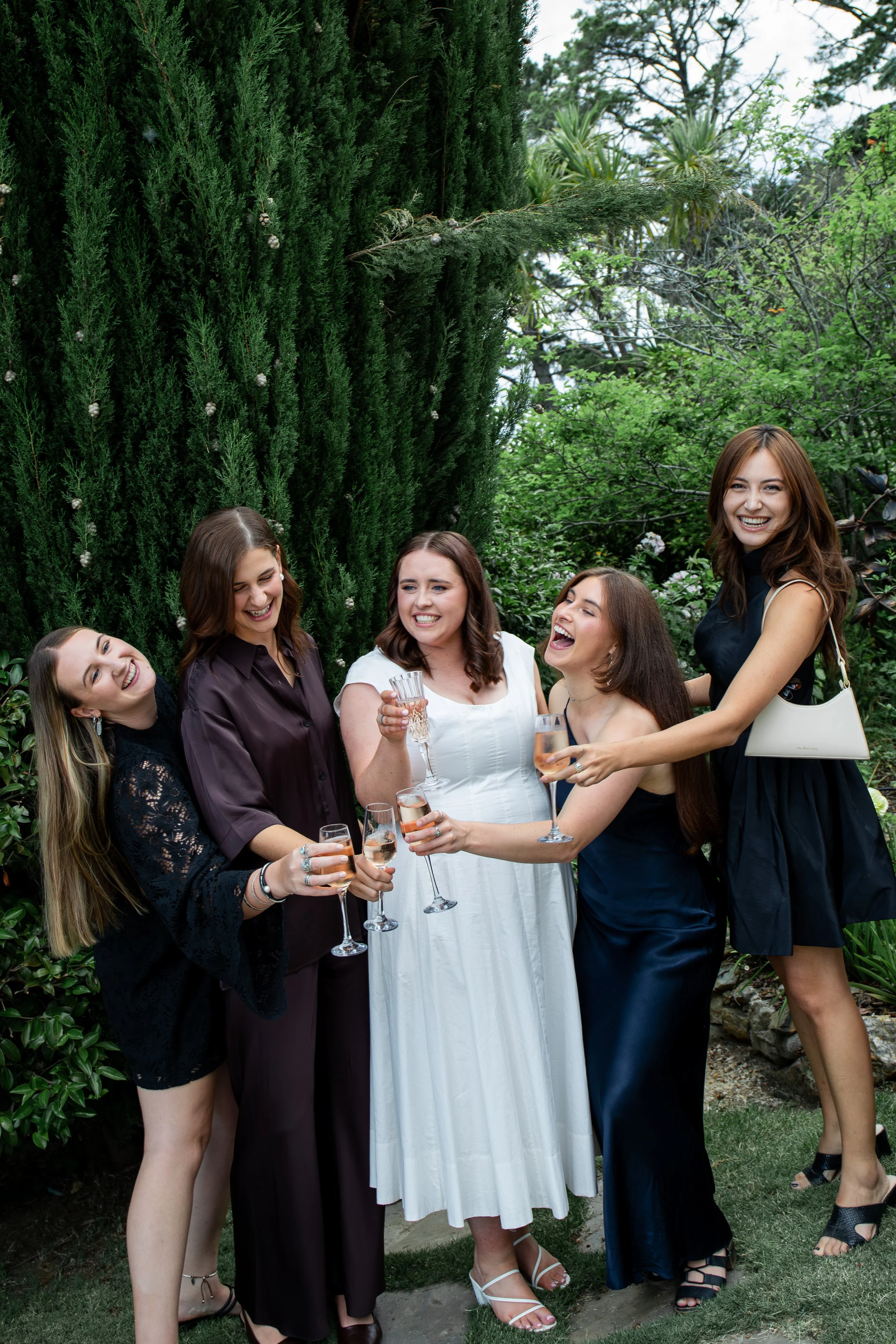 Group of five women celebrating outdoors with drinks, standing on grass with green trees in the background.