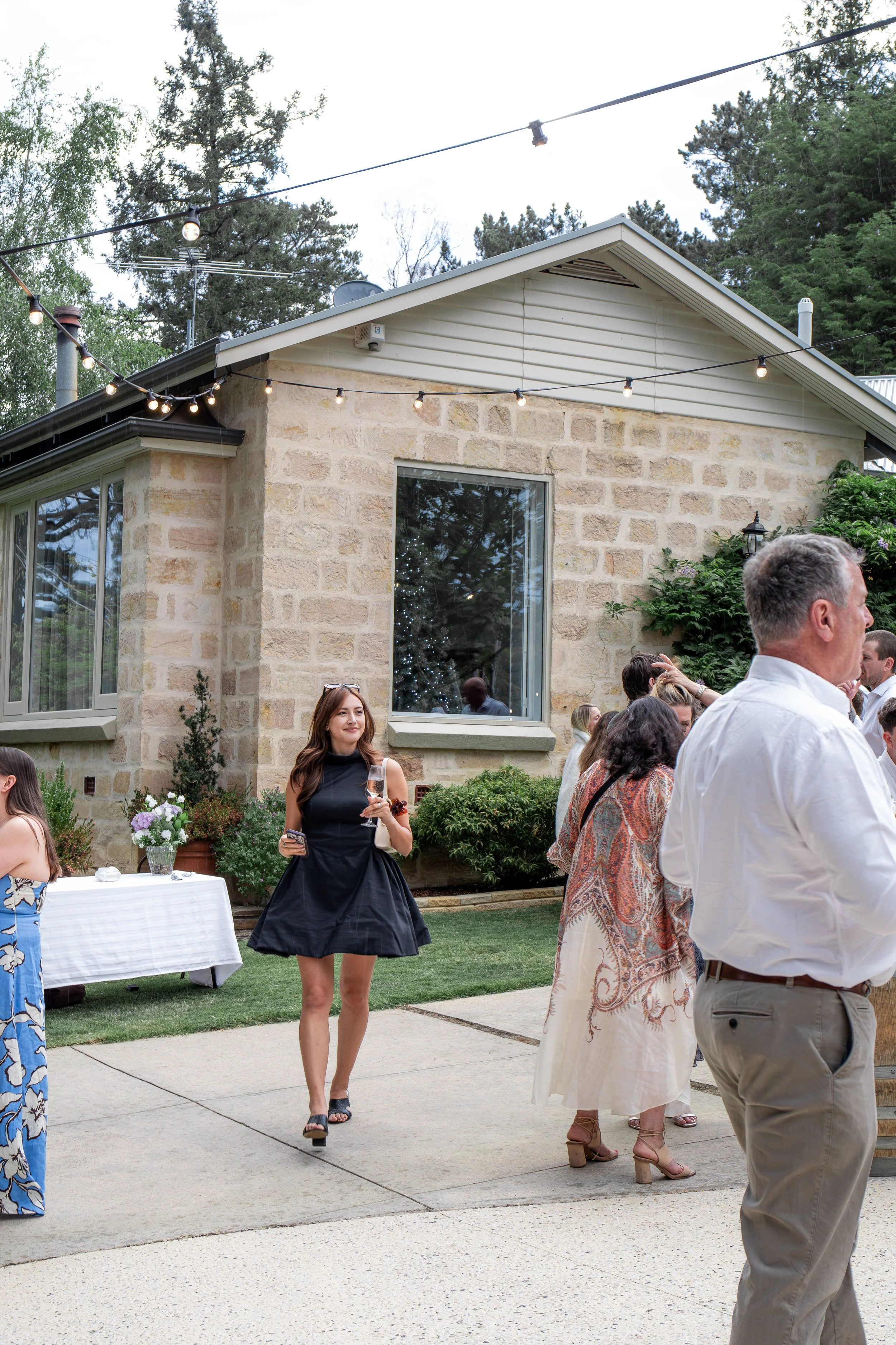 People attending an outdoor social gathering or party on a patio near a stone house with string lights overhead, greenery, and a window reflecting a decorated Christmas tree.