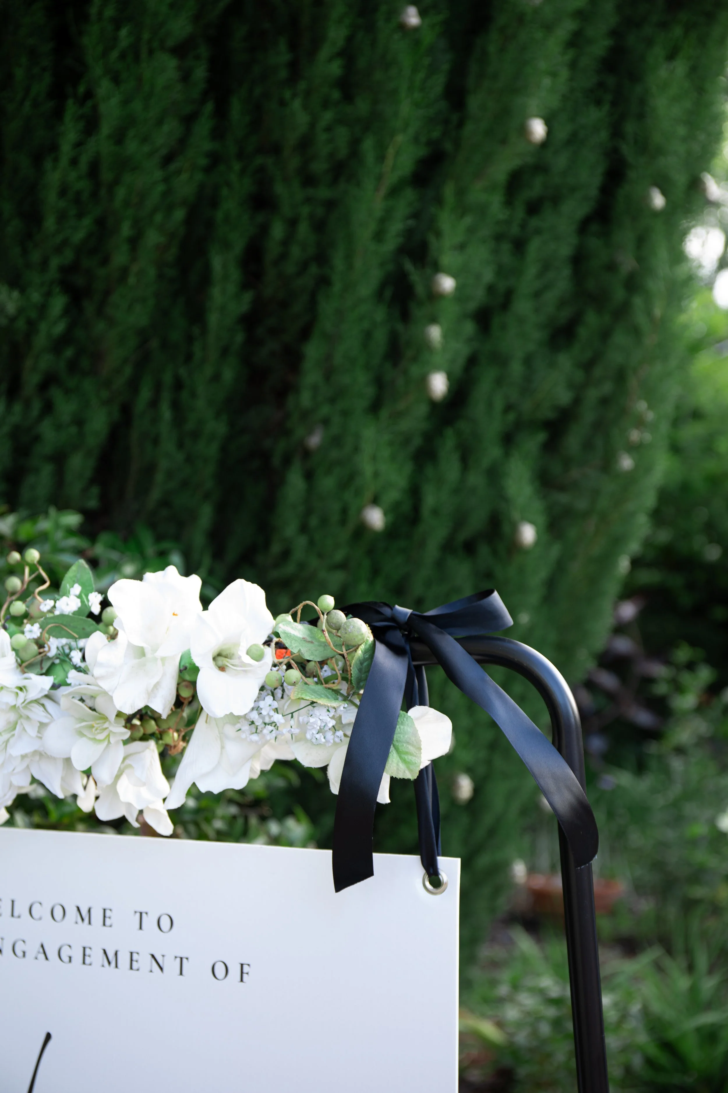 White floral arrangement with black ribbon on a black stand near greenery, with partial text visible that reads 'WELCOME TO ENGAGEMENT OF'.