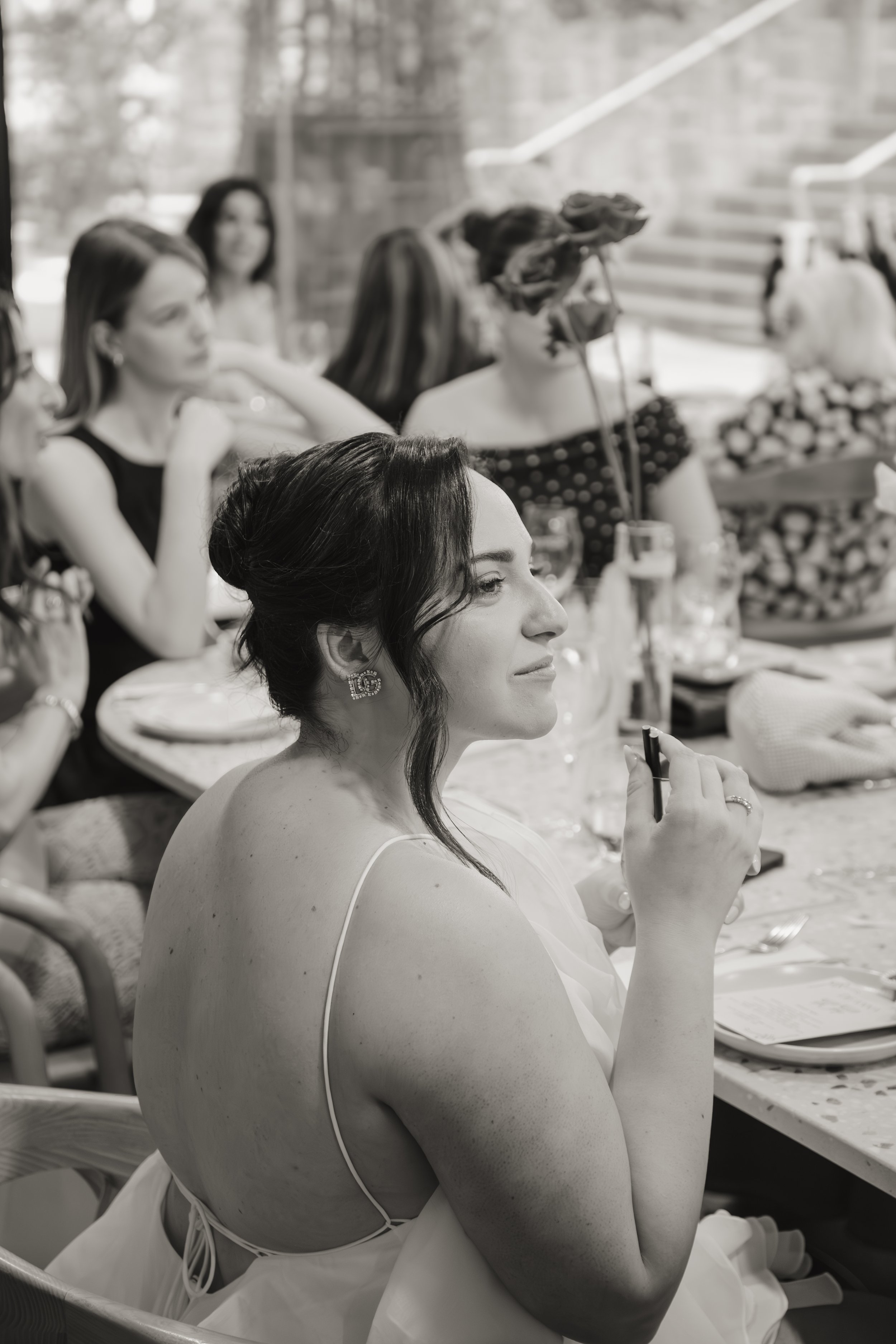 A woman with dark hair styled in an updo, wearing earrings and a sleeveless dress, sitting at a table with others during a celebration, with a person in the background playfully holding a bouquet of flowers over their face.