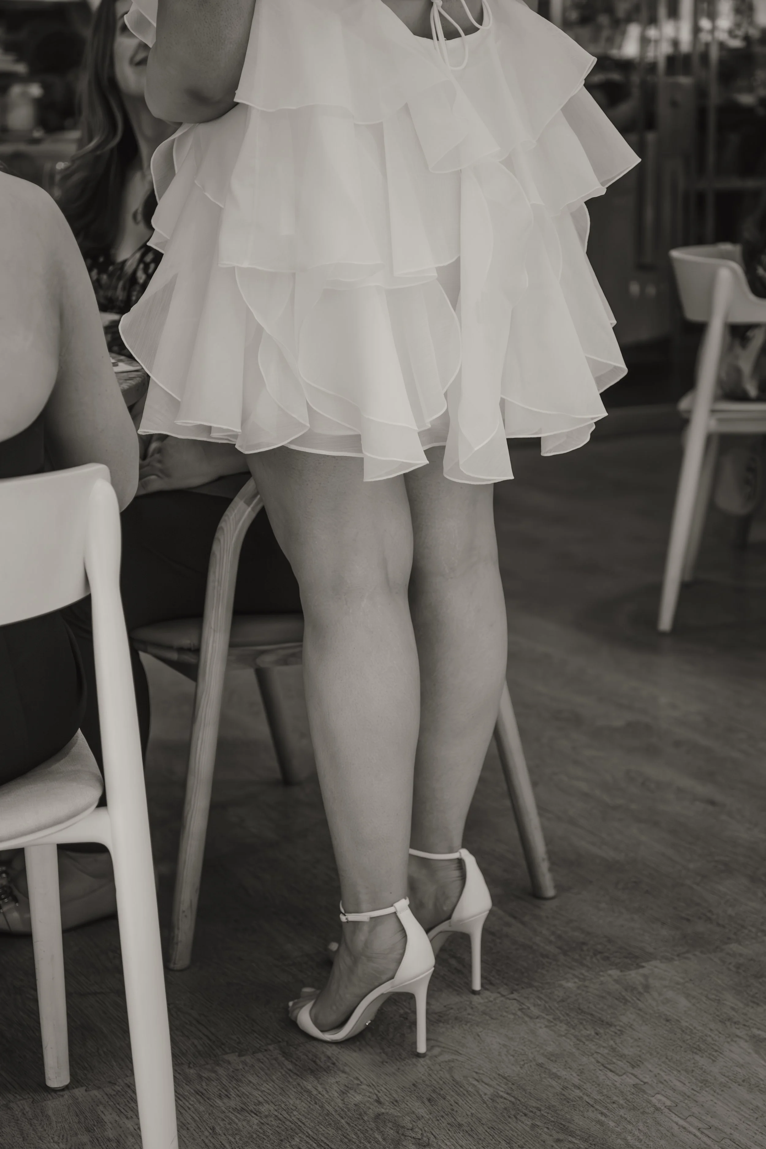 A person in a frilly white dress and white high heels standing next to a seated woman in a restaurant.