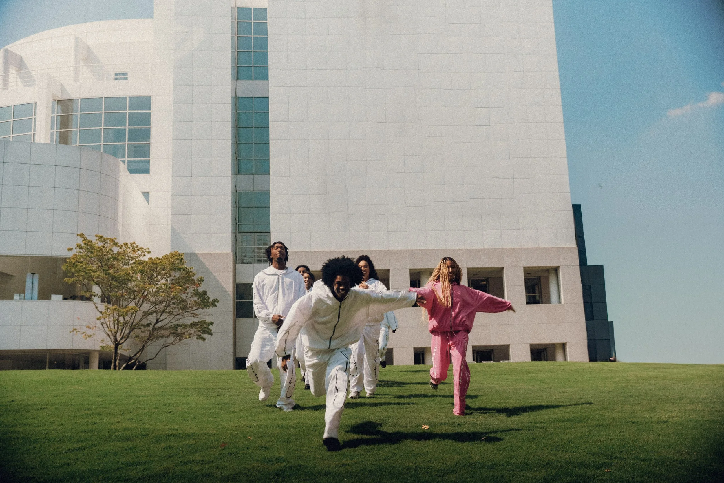 Group of young people running on a grassy lawn in front of a modern white building, with clear blue sky in the background.