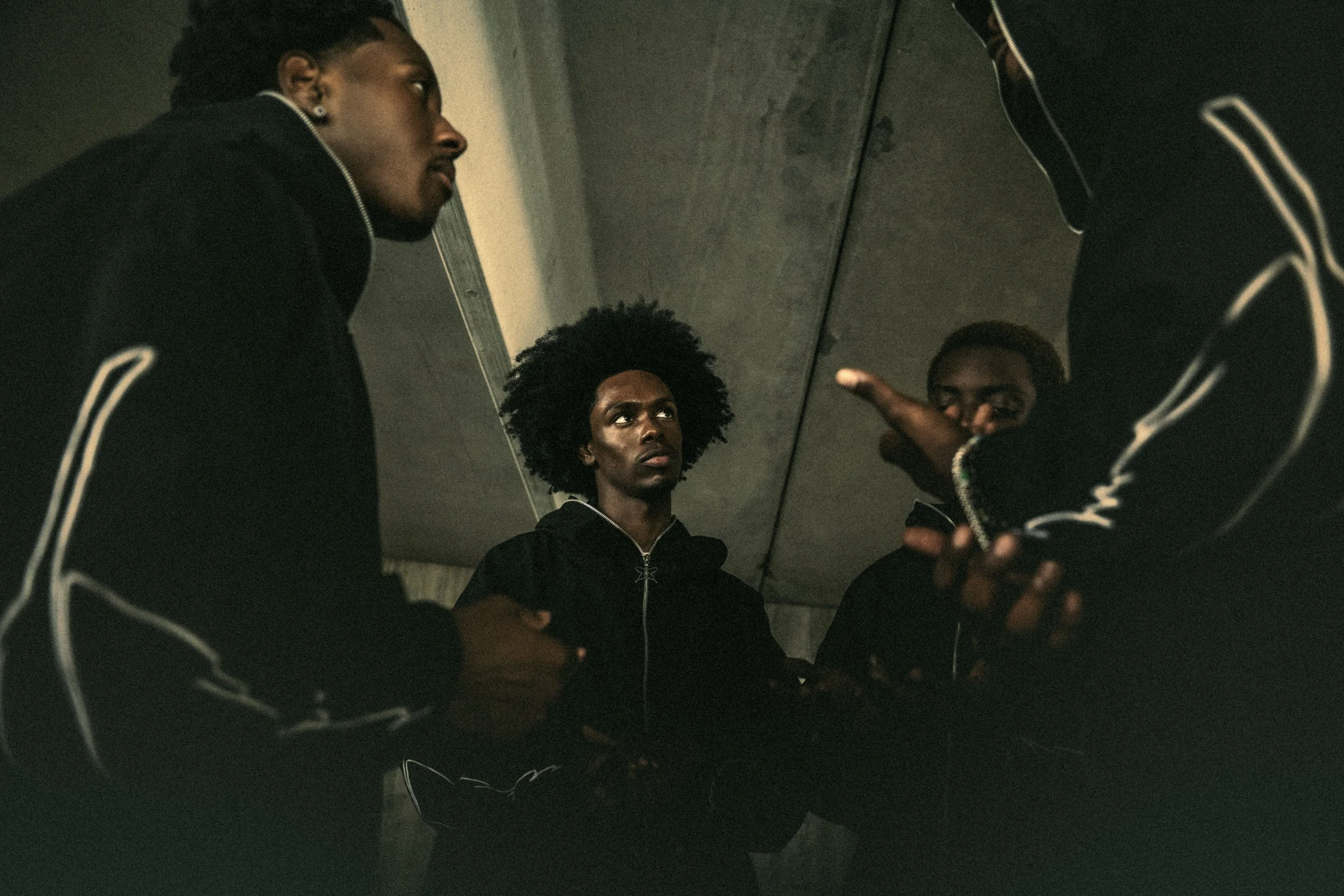 Four young adults in black jackets with white piping sitting and talking under a concrete bridge.