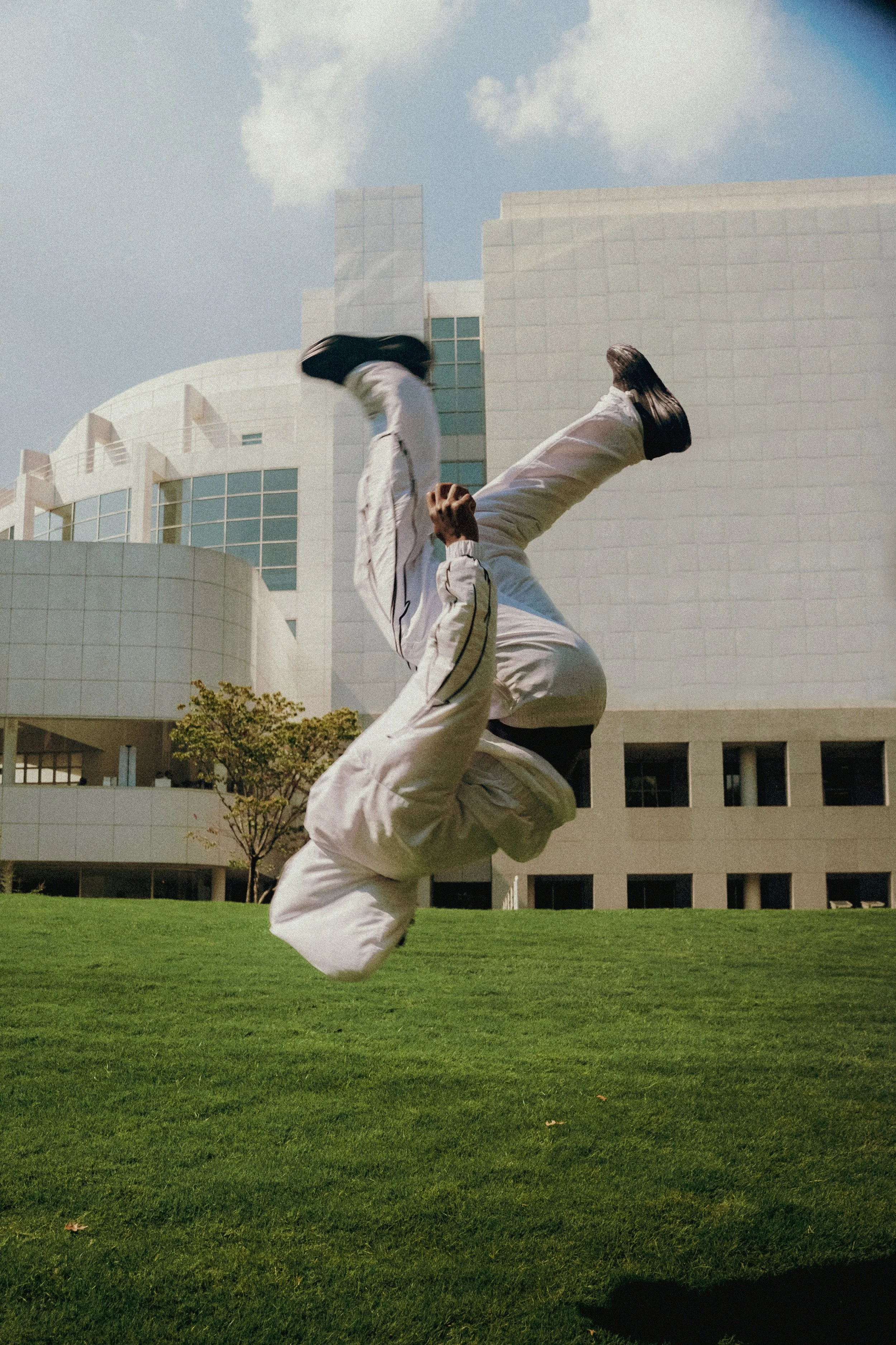 Person performing a backflip outdoors in front of a modern building with green grass and blue sky.
