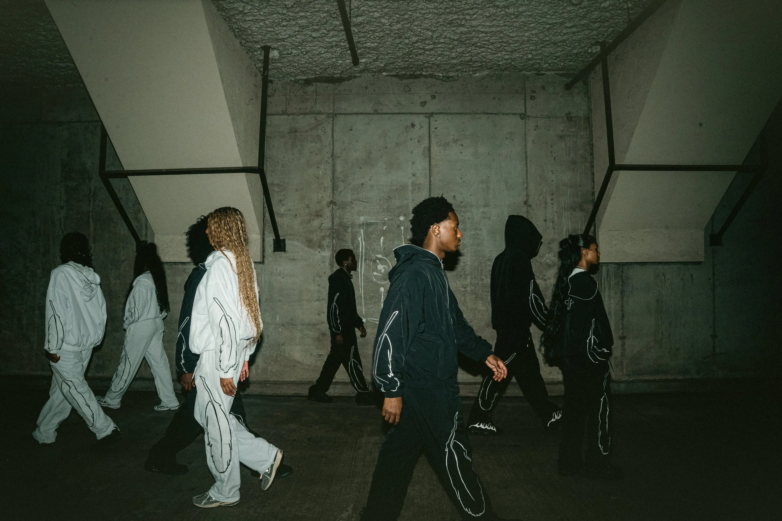 A group of six people walking in a line underground with concrete walls and ceiling, wearing matching black and white tracksuits with abstract line designs.
