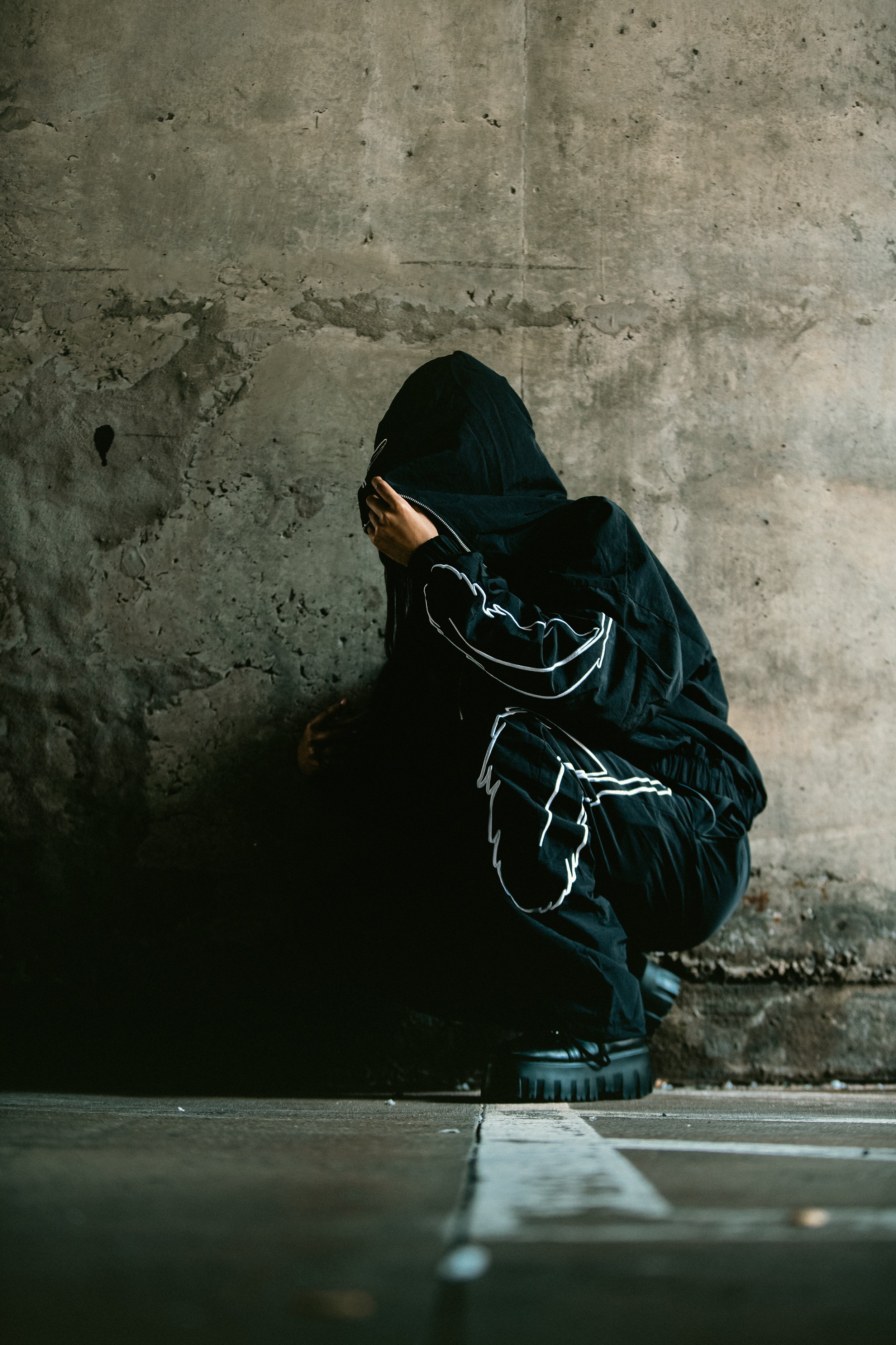 A person dressed in black crouching against a concrete wall, with their face covered by their hand.