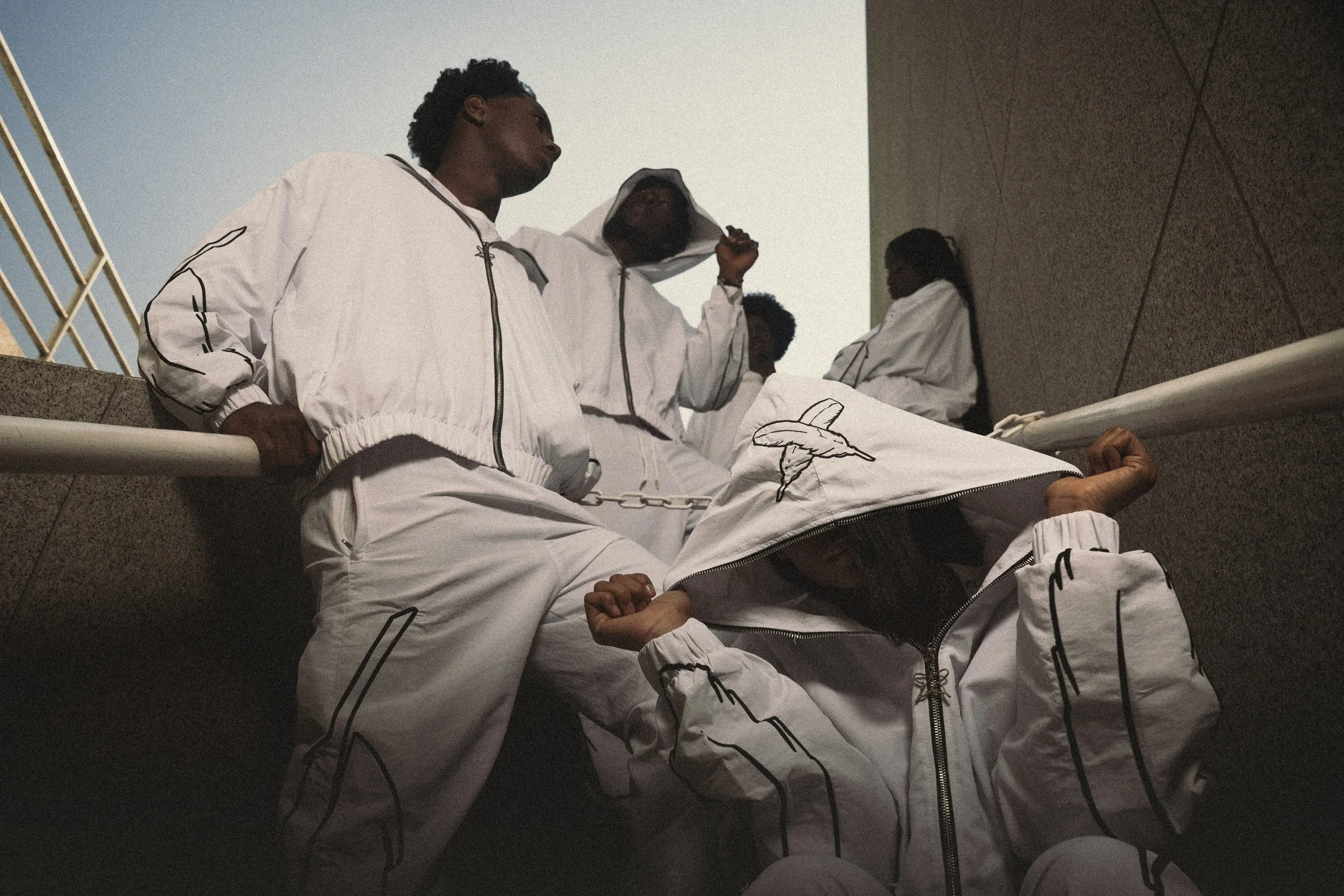 A group of young people in matching white outfits with black accents, some wearing hoodies, gathered on an outdoor staircase, with one person sitting and others standing around.