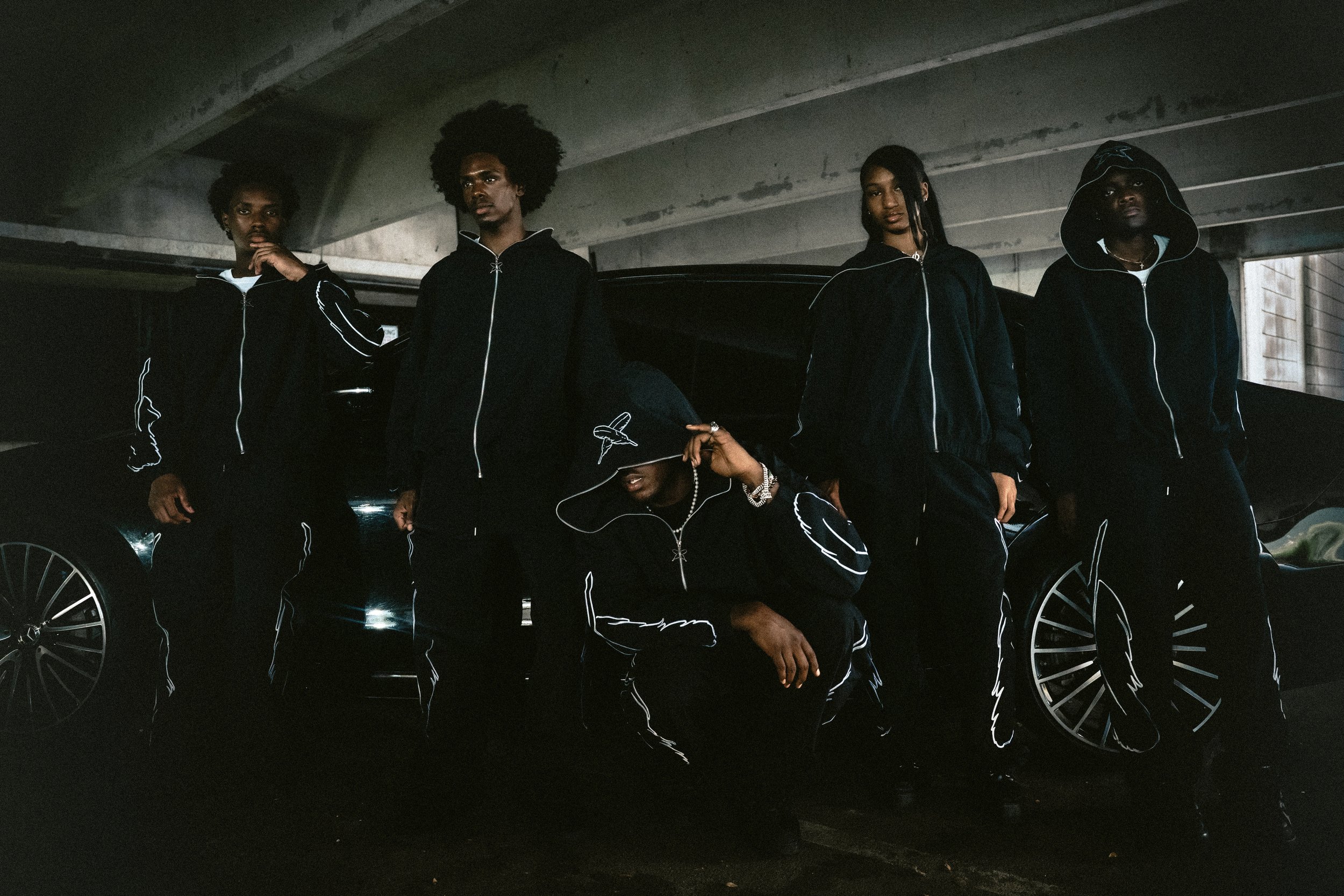 Group of five young men dressed in black hoodies and track pants, standing and kneeling in a parking garage with a black car behind them.