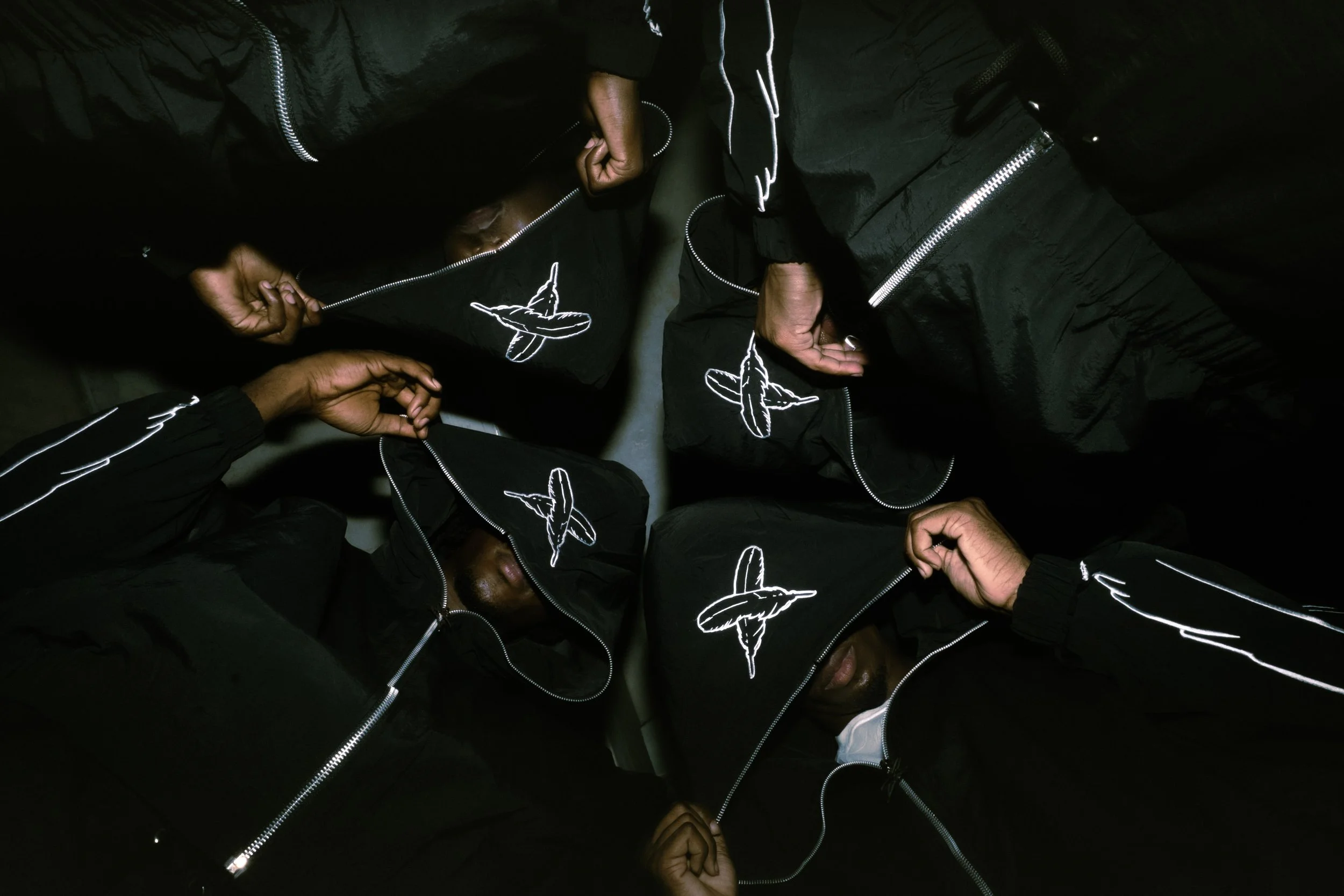 Four people in black hooded jackets with white embroidery of a plane on the hood pulling down their hoods in a dark room.