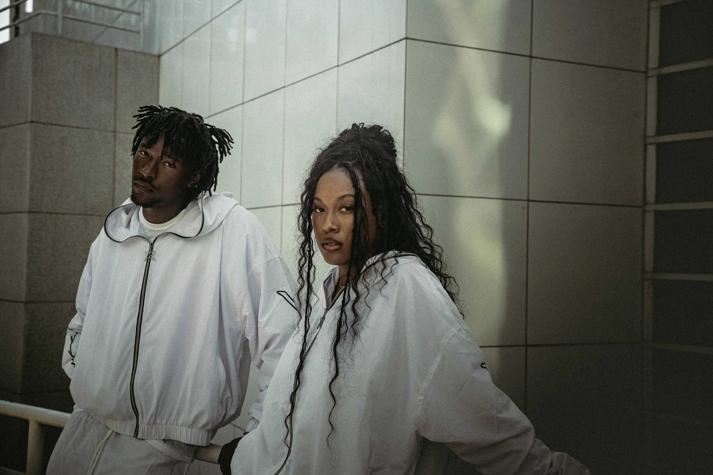 Two young adults, a man and a woman, wearing white jackets, standing against a modern tiled wall in an urban setting, looking at the camera with confident expressions.