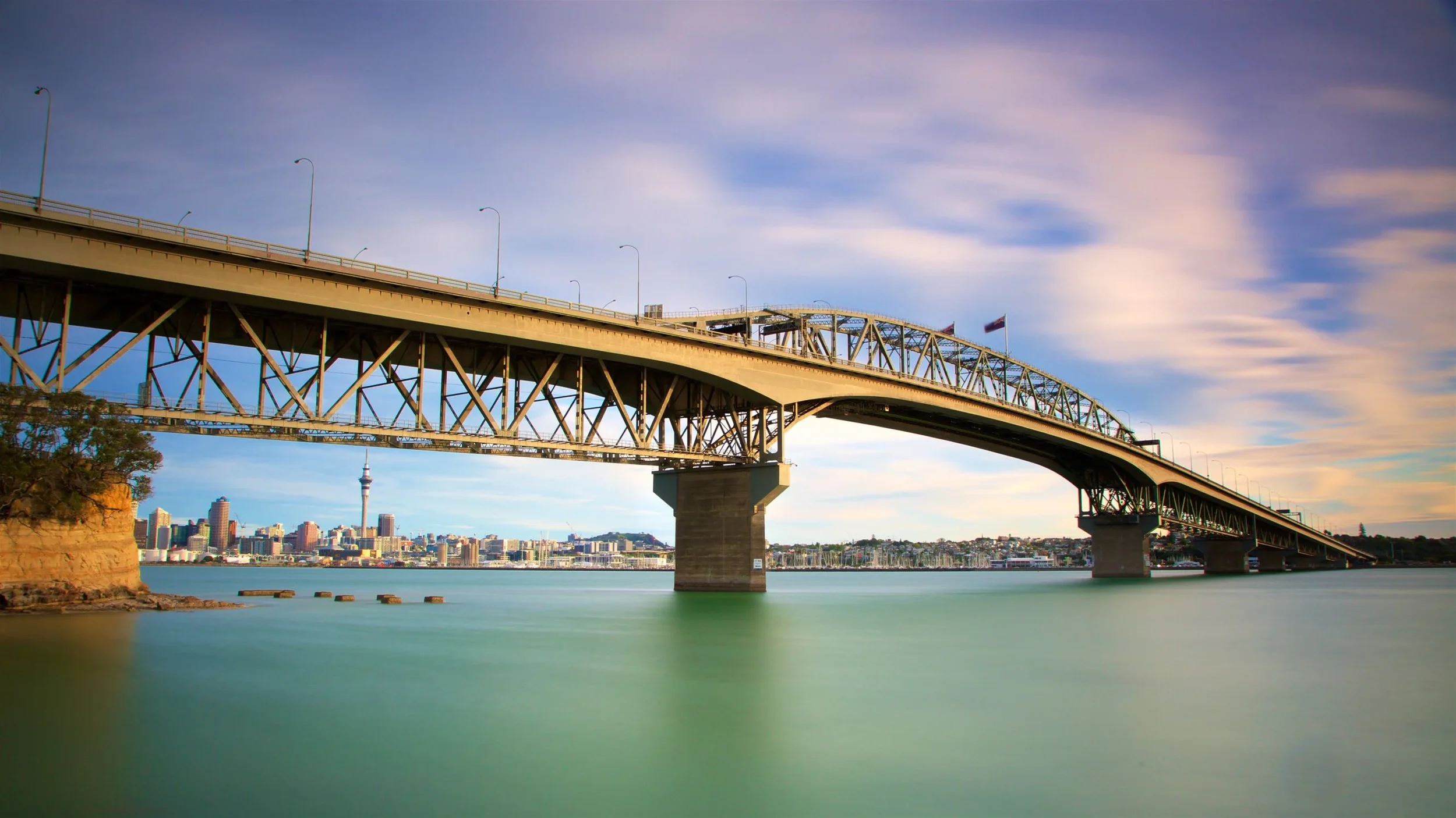 Puente sobre agua con ciudad y torre en el fondo, cielo con nubes y línea de horizonte.