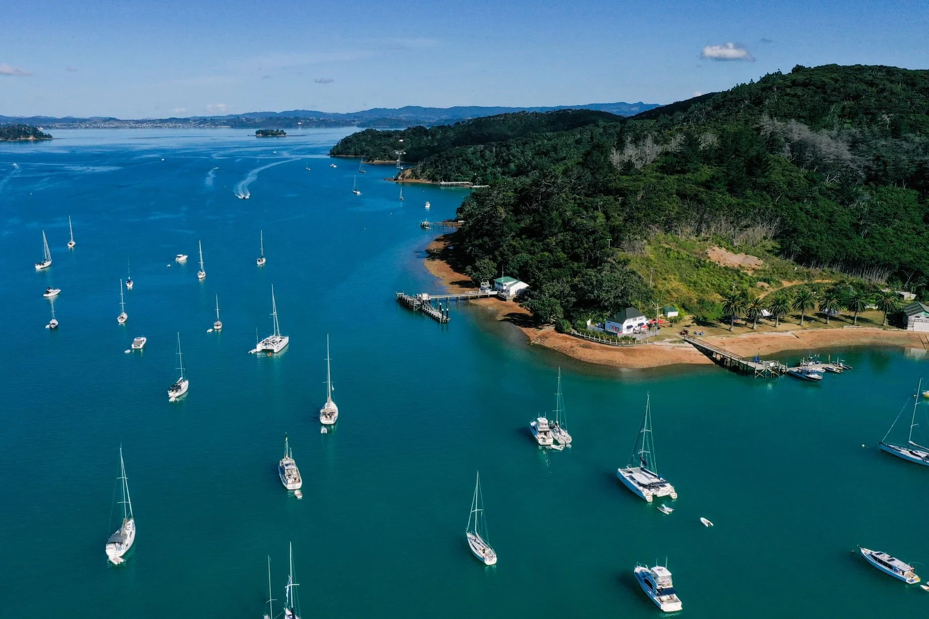 Vista aérea de un puerto con varios barcos y yates anclados en aguas tranquilas, rodeado de colinas verdes y bosques, con un pequeño muelle y casas en la orilla.