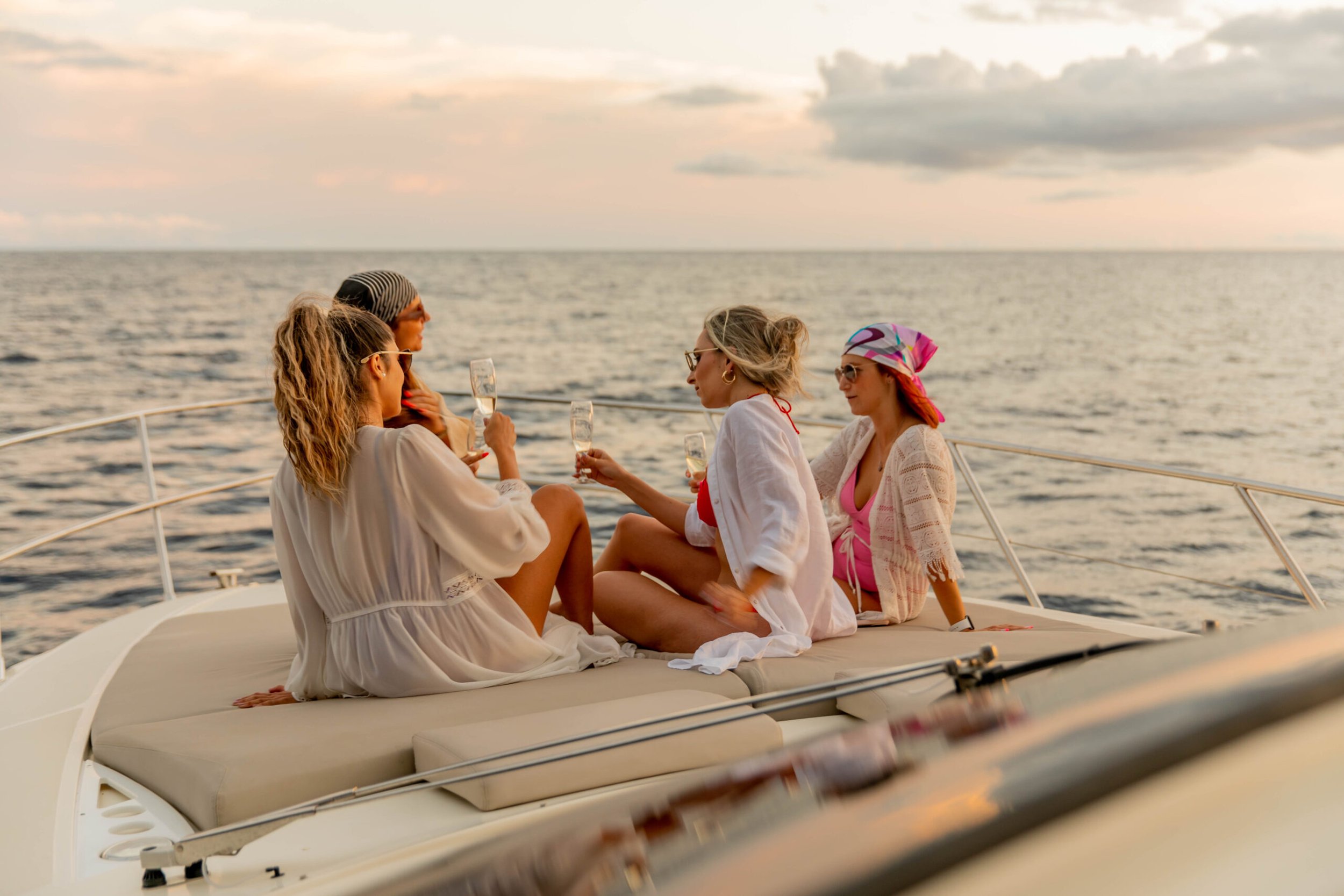 Grupo de cuatro mujeres en un yate, disfrutando una copa de champán al atardecer en el mar.
