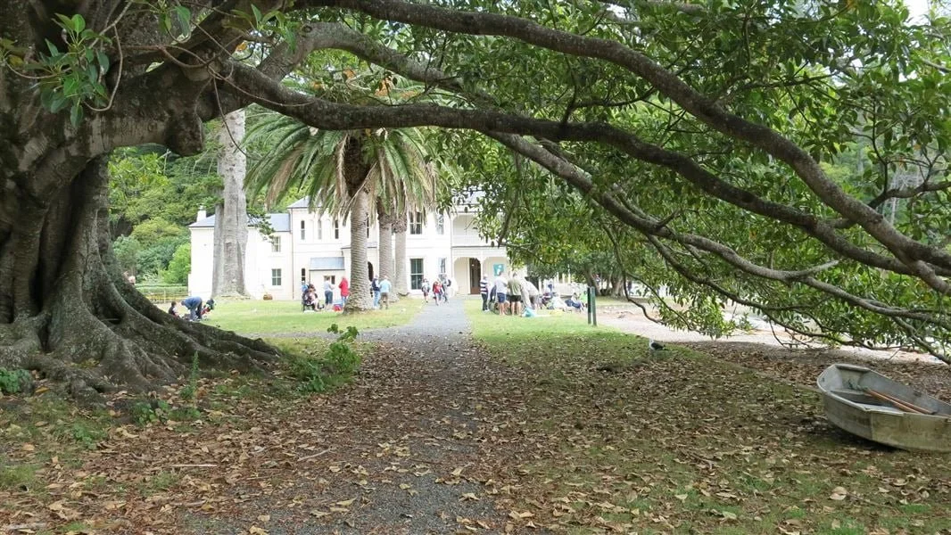 Camino entre árboles que conduce a una casa grande, con gente reunida en el fondo, y un bote en el lado derecho.