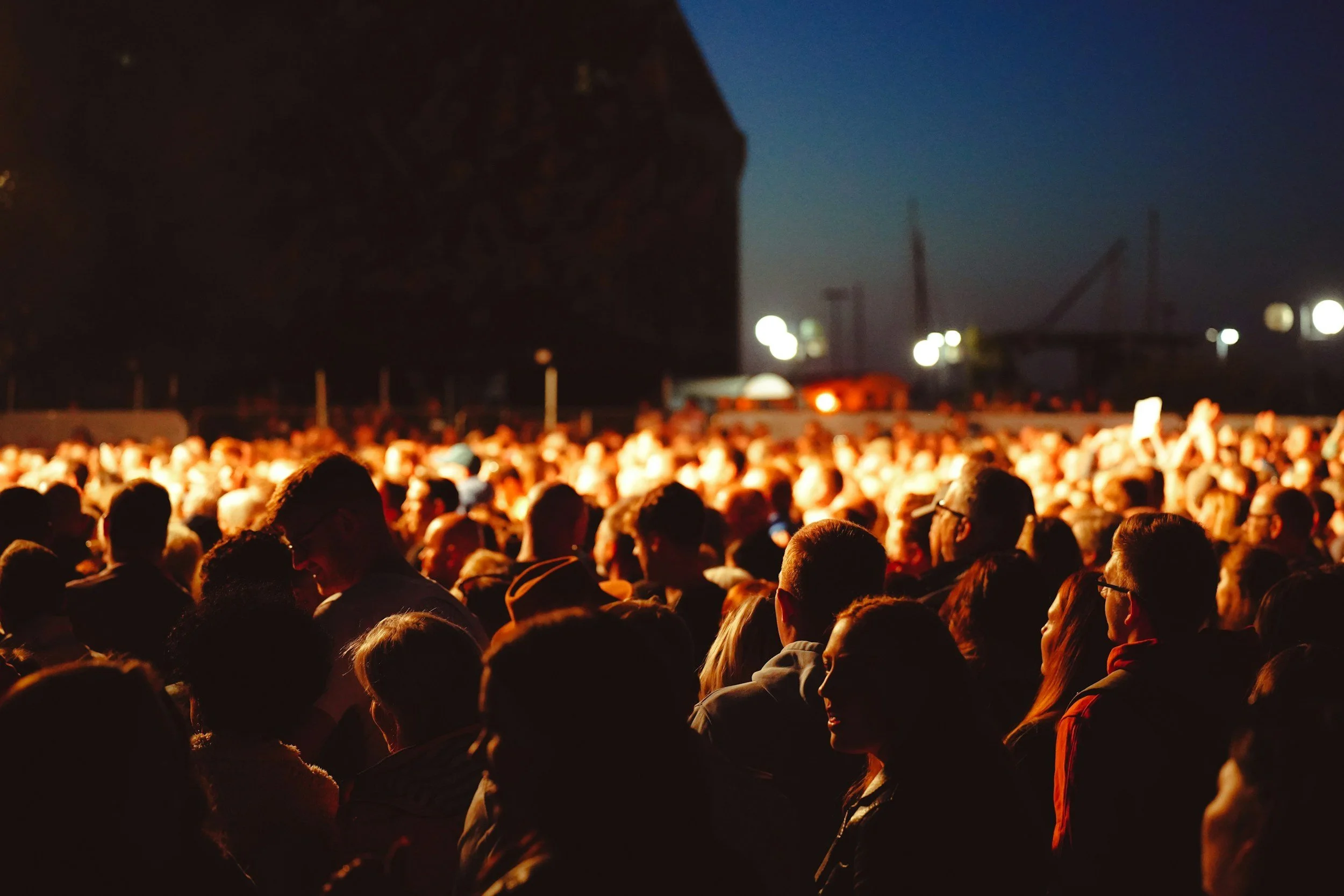 Crowd of people gathered outdoors at night, illuminated by warm lights, with a dark sky and distant structures visible in the background.