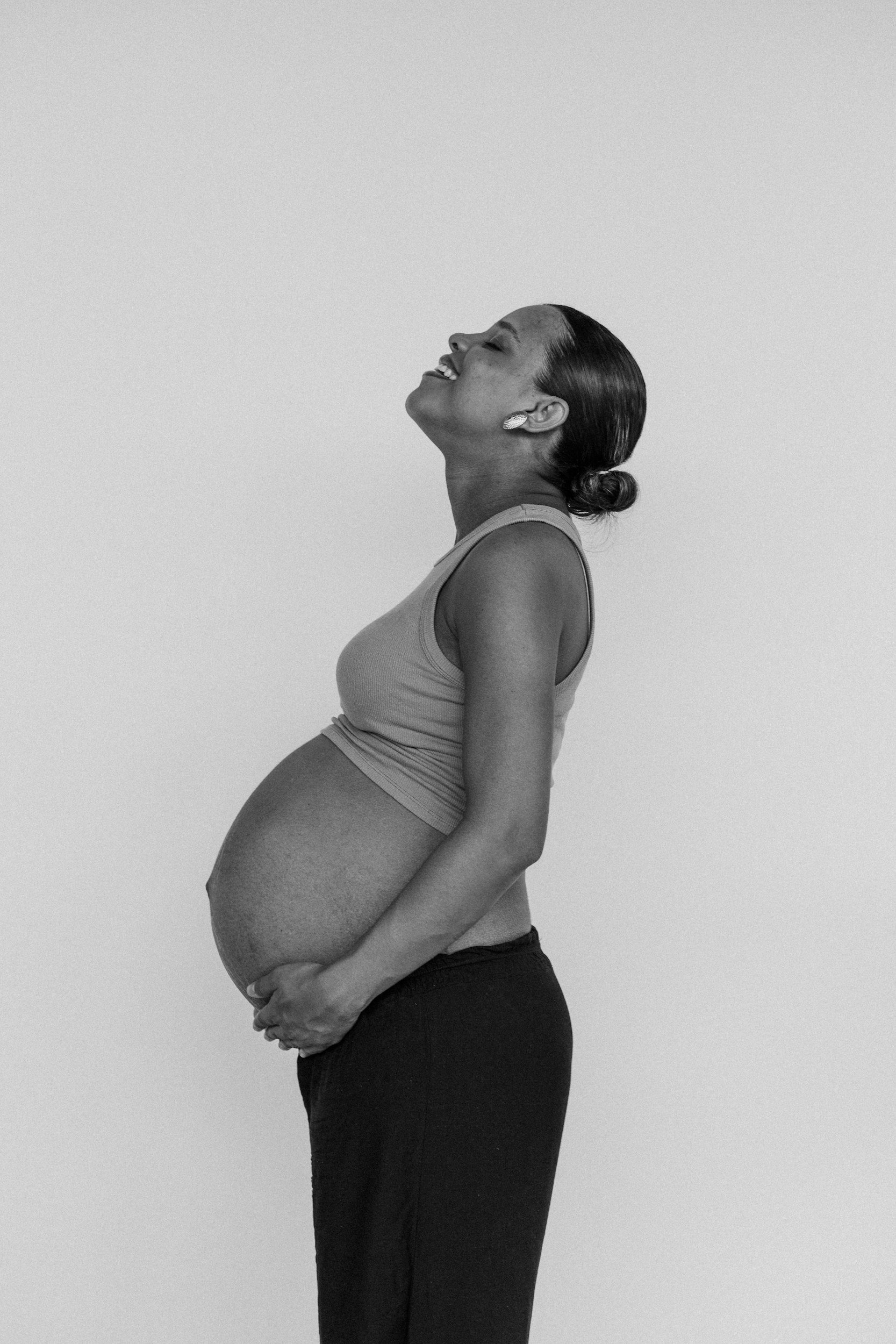 Black and white photograph of a pregnant woman in profile, smiling and looking upward, wearing a tank top and dark pants, with her hair tied back and one hand on her belly.