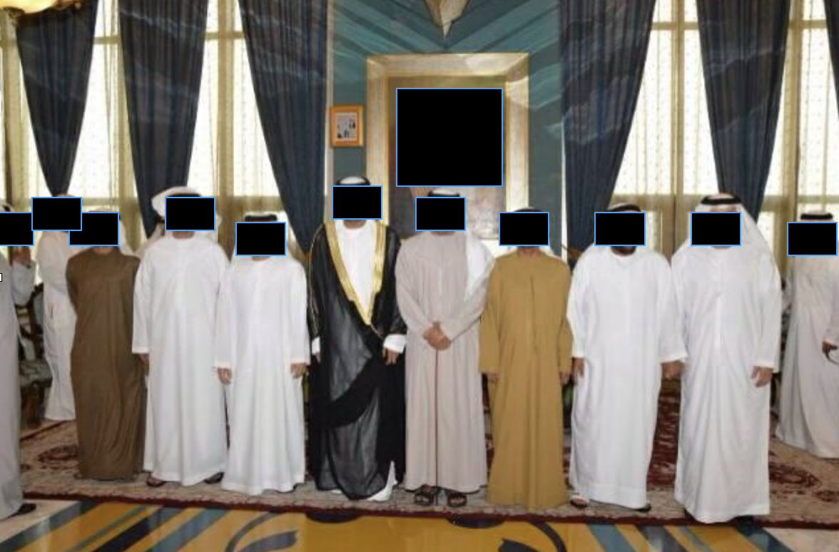 Group of people standing in a room with draped curtains and tall windows, some wearing traditional Middle Eastern clothing, posing for a photo.