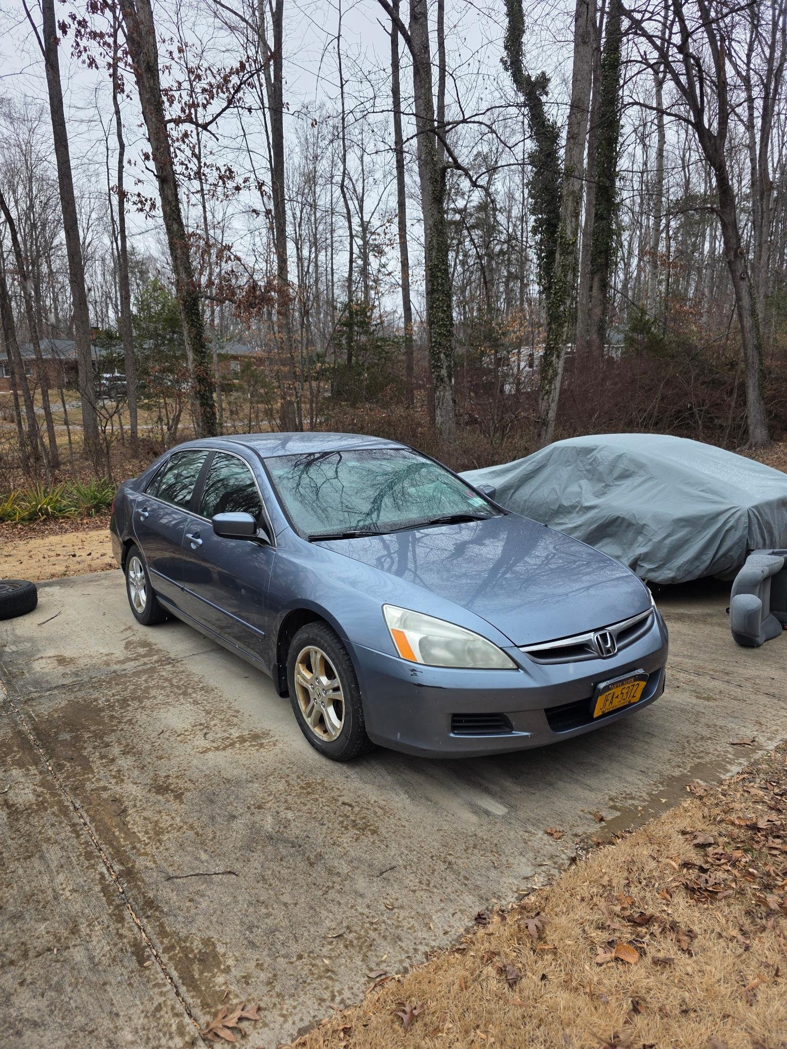 A blue Honda sedan parked on a concrete driveway surrounded by leafless trees and a forest in the background. The car has some scratches and wears gold-colored rims, and there is a covered vehicle nearby.