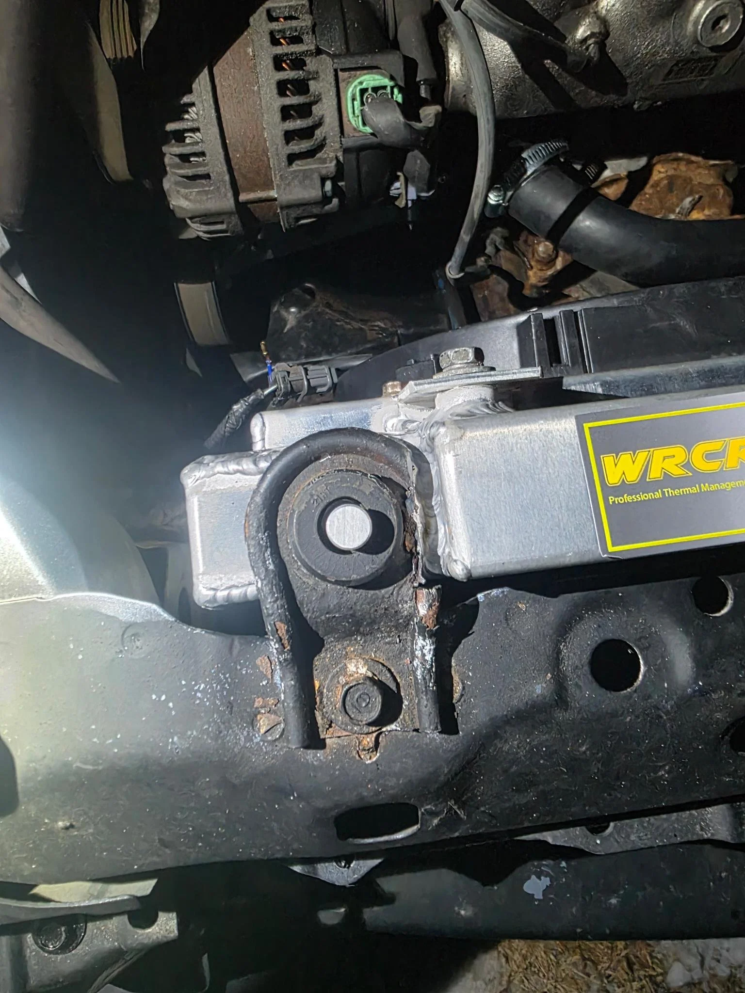 Close-up of a modified car radiatormount, connected to an aluminum component labeled WRCF, part of the vehicle's thermal management system.