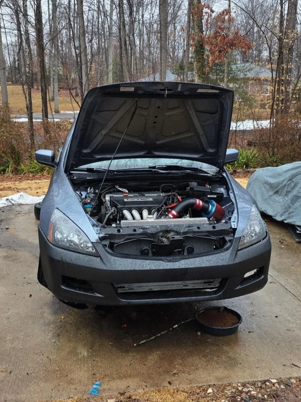 A black car with its hood open, exposing the engine bay with aftermarket parts like a red air filter and intercooler piping, parked outdoors on a driveway with trees and houses in the background.