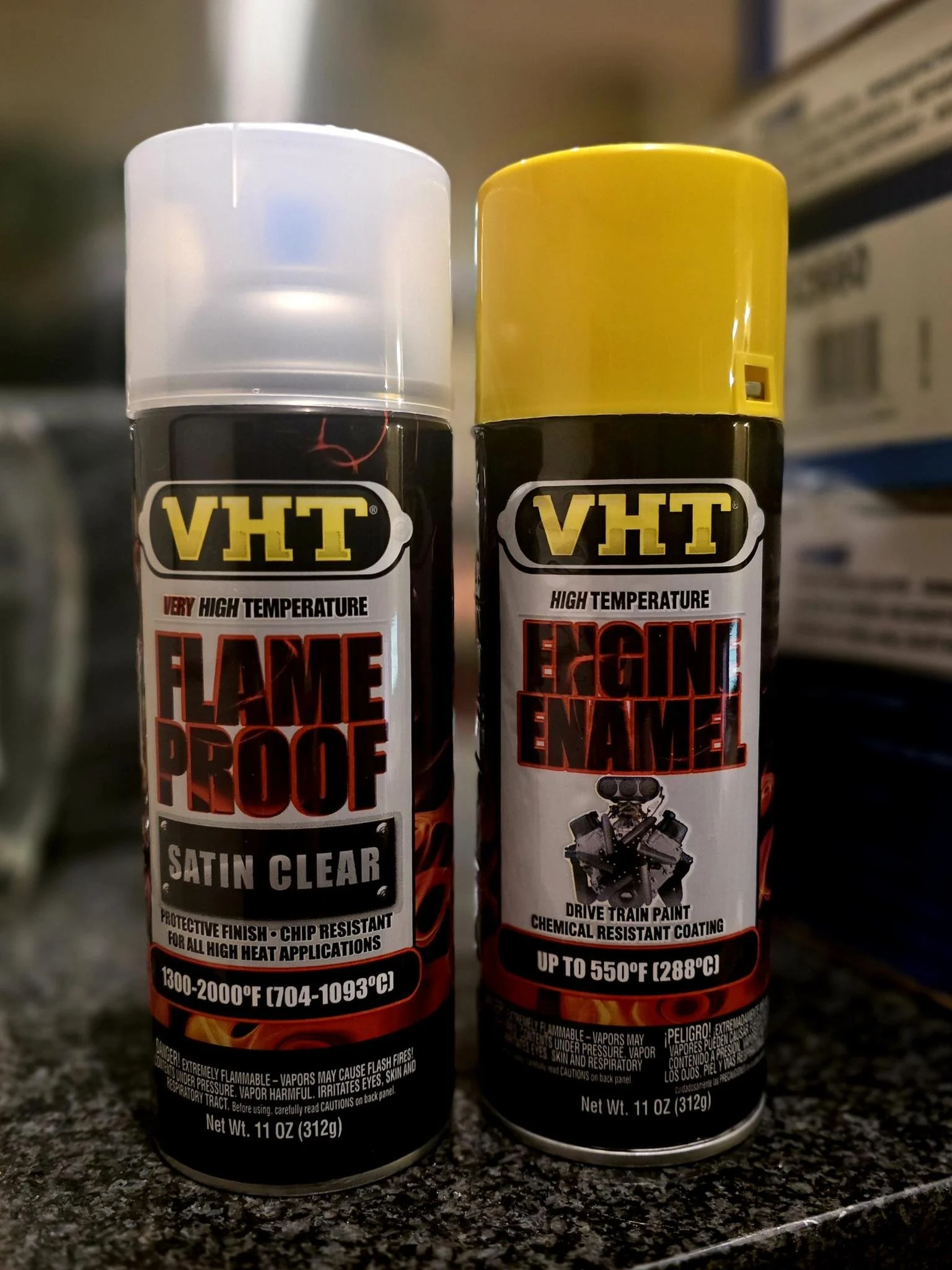 Two cans of automotive high-temperature spray paint with black and yellow caps, labeled "VHT Flame Proof Satin Clear" and "VHT Reign Enamel," placed on a dark countertop in a store.