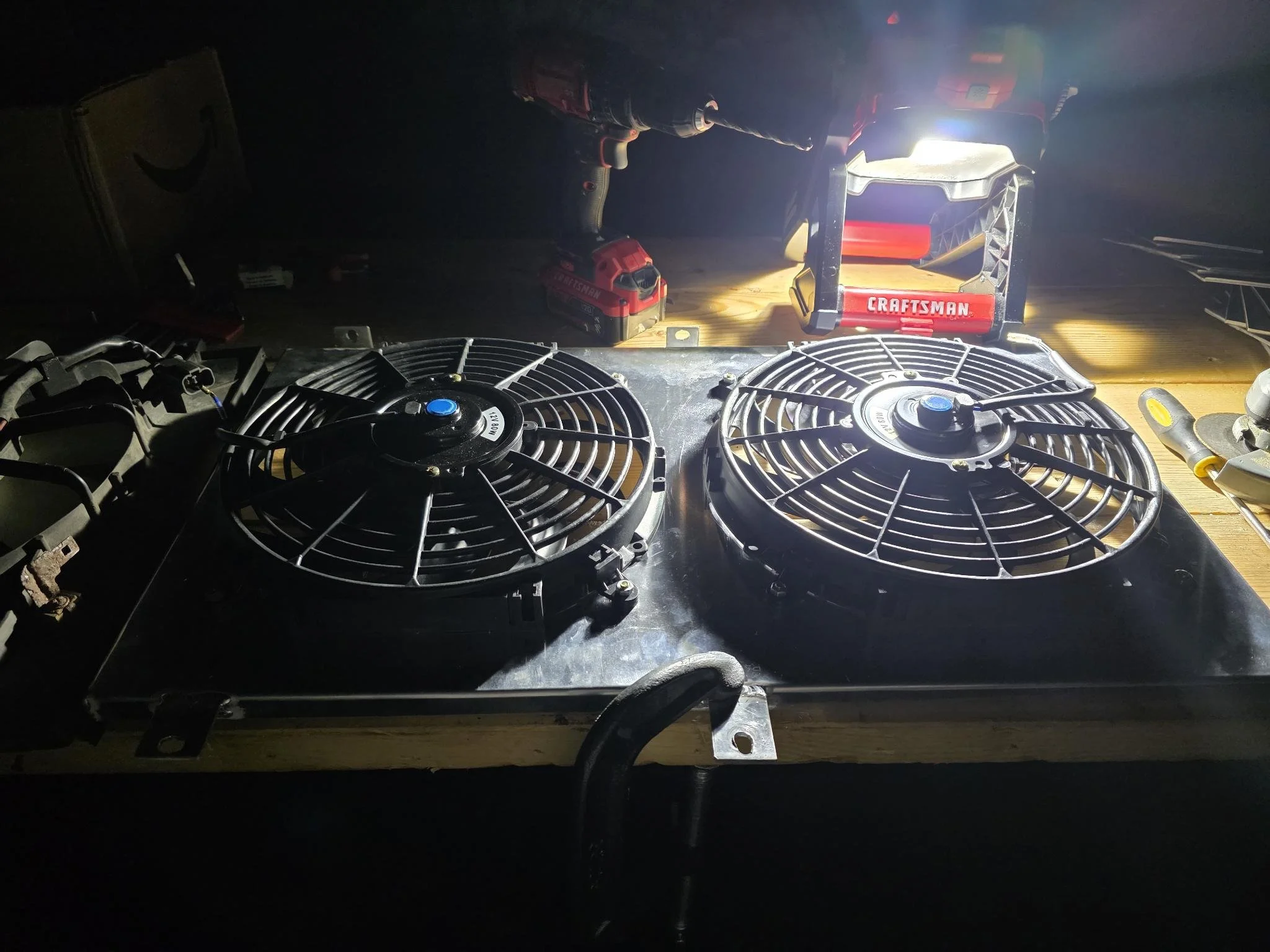 Two electric radiator cooling fans on a workbench illuminated by a bright light. Various tools and equipment, including a red tool box labeled 'CRAFTSMAN' and a cordless drill, are visible around the work area.