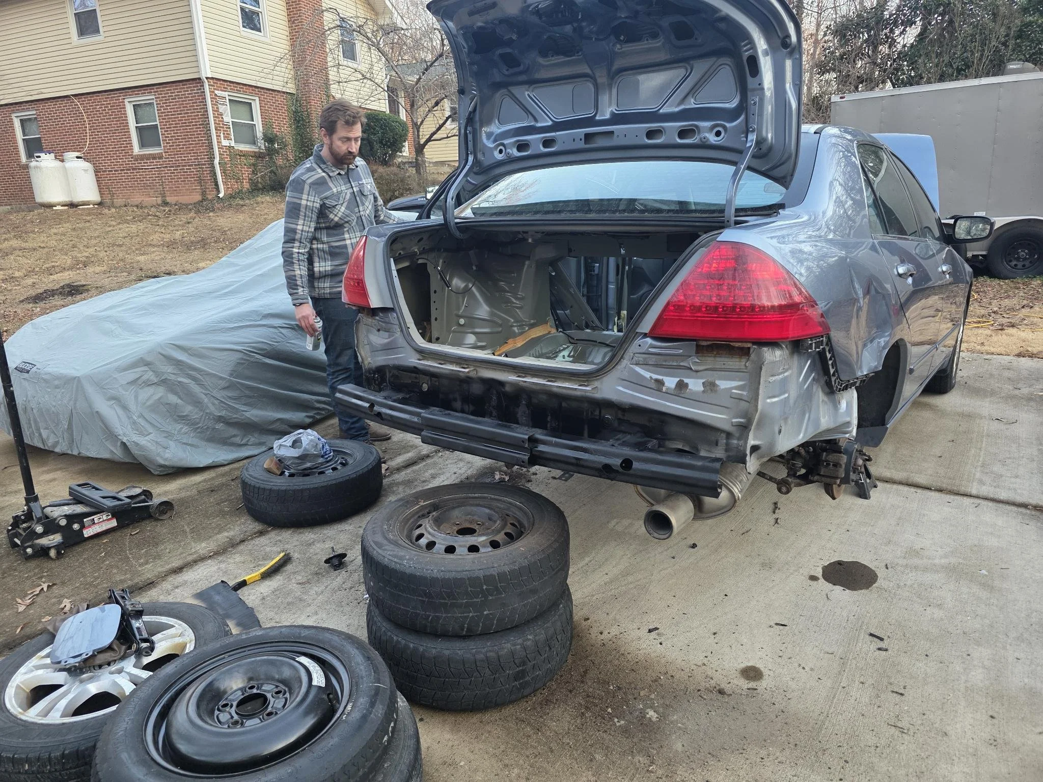 Man working on a  2007 Honda Accord with the rear bumper removed, tires and tools on the ground, and a covered car in the background.