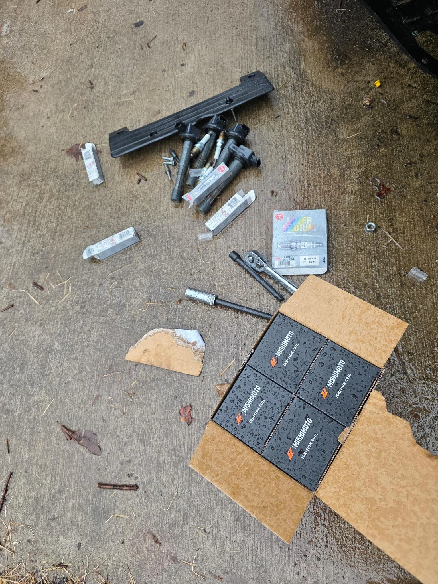 Car parts and tools scattered on a wet concrete driveway, including spark plugs, a box of automotive parts, and various small hardware items.