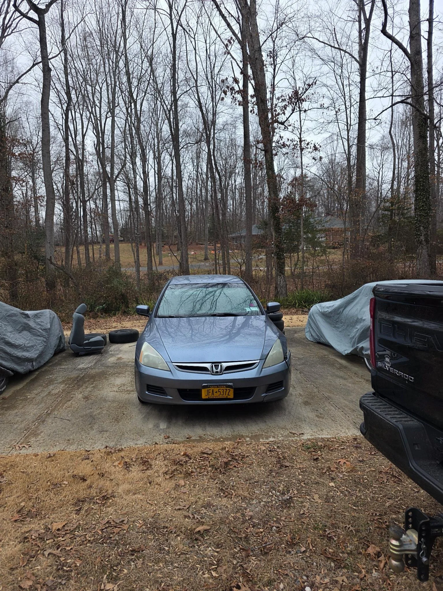 A front view of a silver Honda car parked in an outdoor area with leafless trees in the background. Two covered vehicles are on either side, and an automotive seat and a tire are visible near the left side of the Honda. The ground is partly covered w