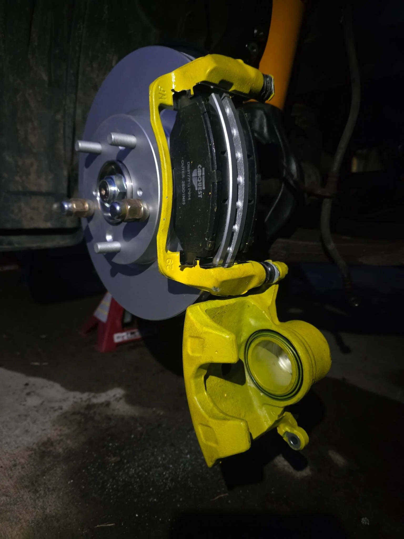 A car's brake assembly with yellow caliper, brake disc, and brake pads mounted on the wheel hub, with various bolts and a protective cover visible.