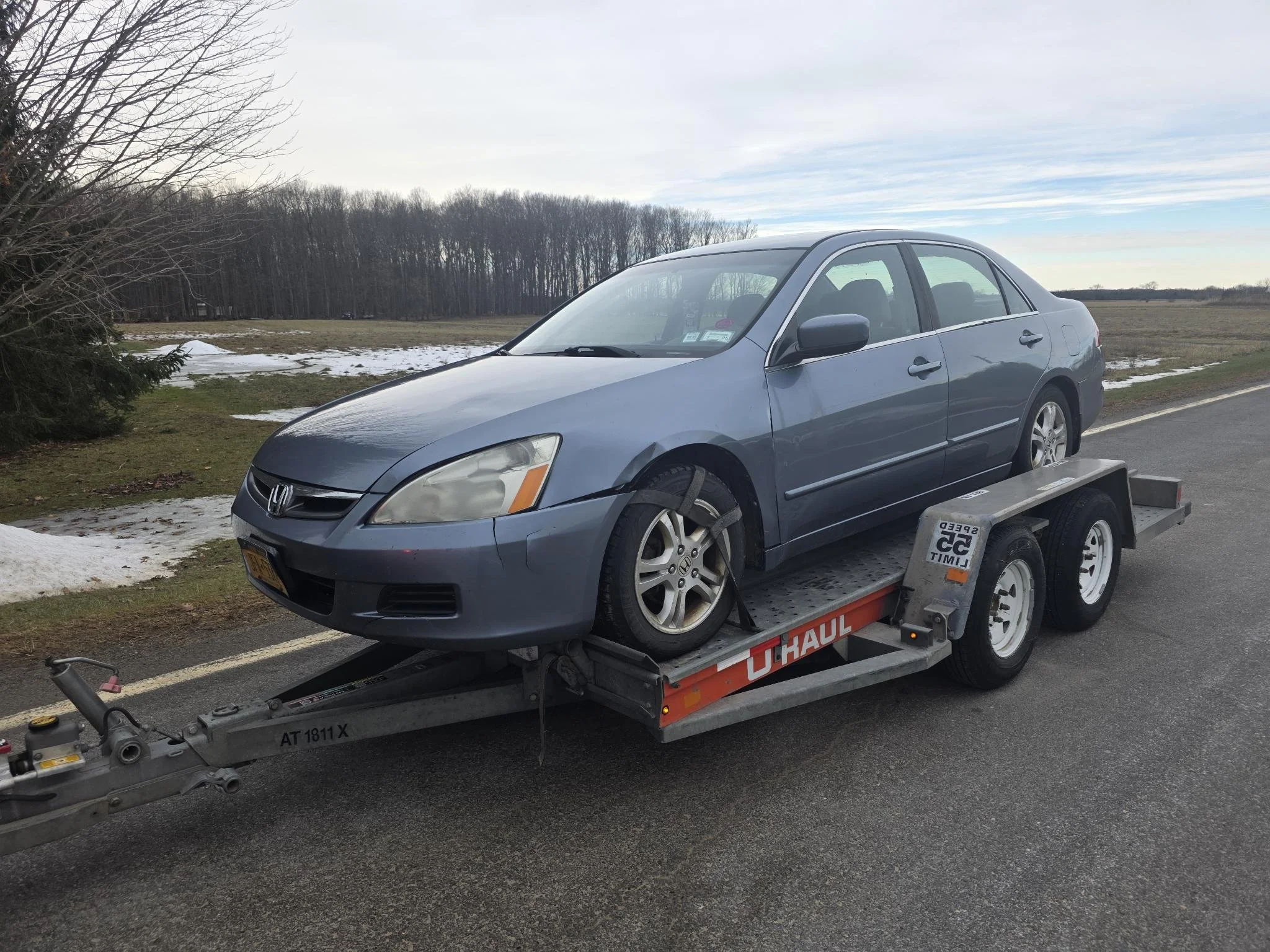 A blue Honda sedan on a U-Haul flatbed trailer parked on the side of a rural road with some snow on the ground and leafless trees in the background.