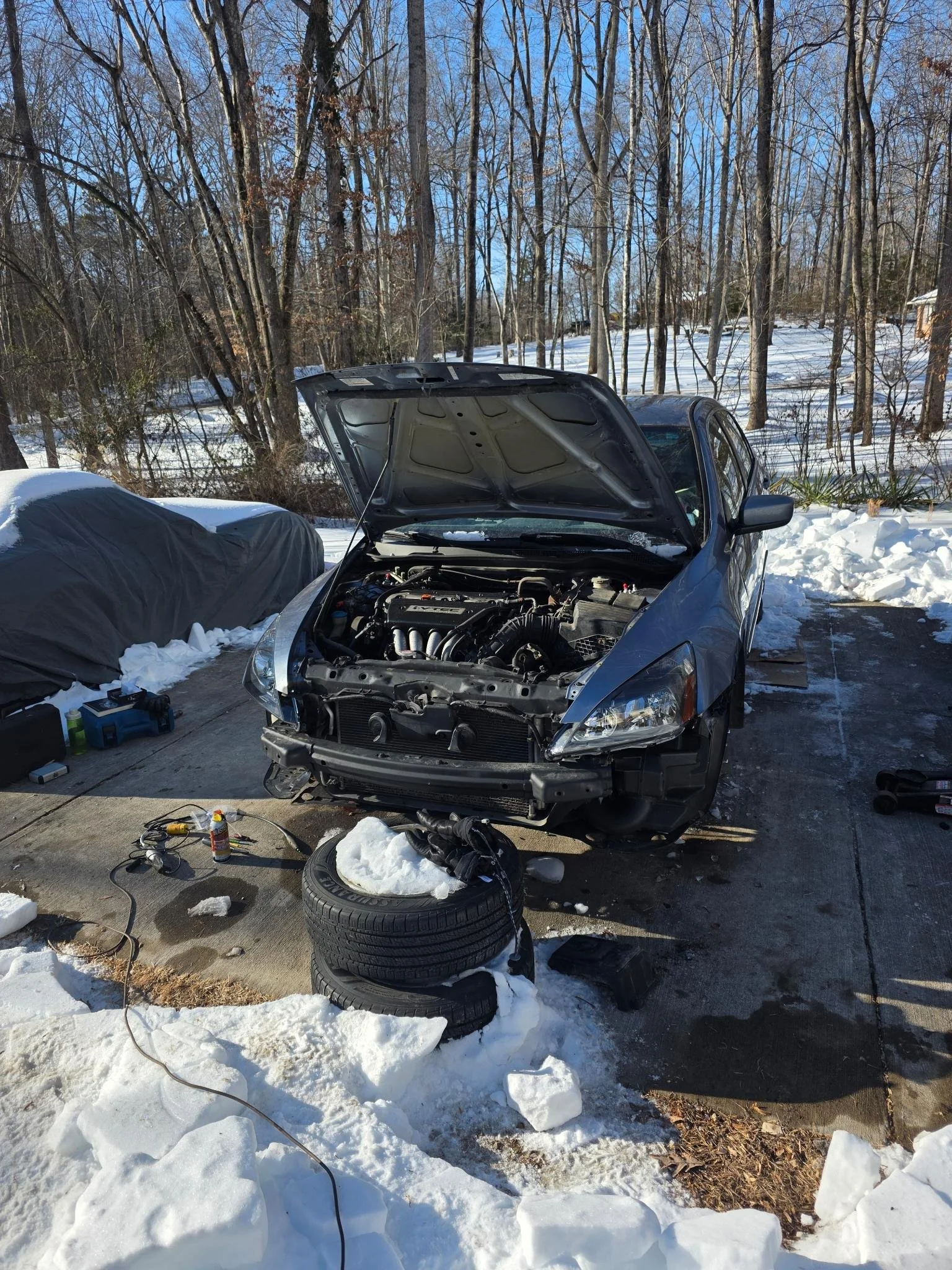 A 2007 Honda Accord hood open, parked on a driveway in winter, undergoing repairs. Tools and spare tires are next to the car, with snow around it and a wooded area in the background.