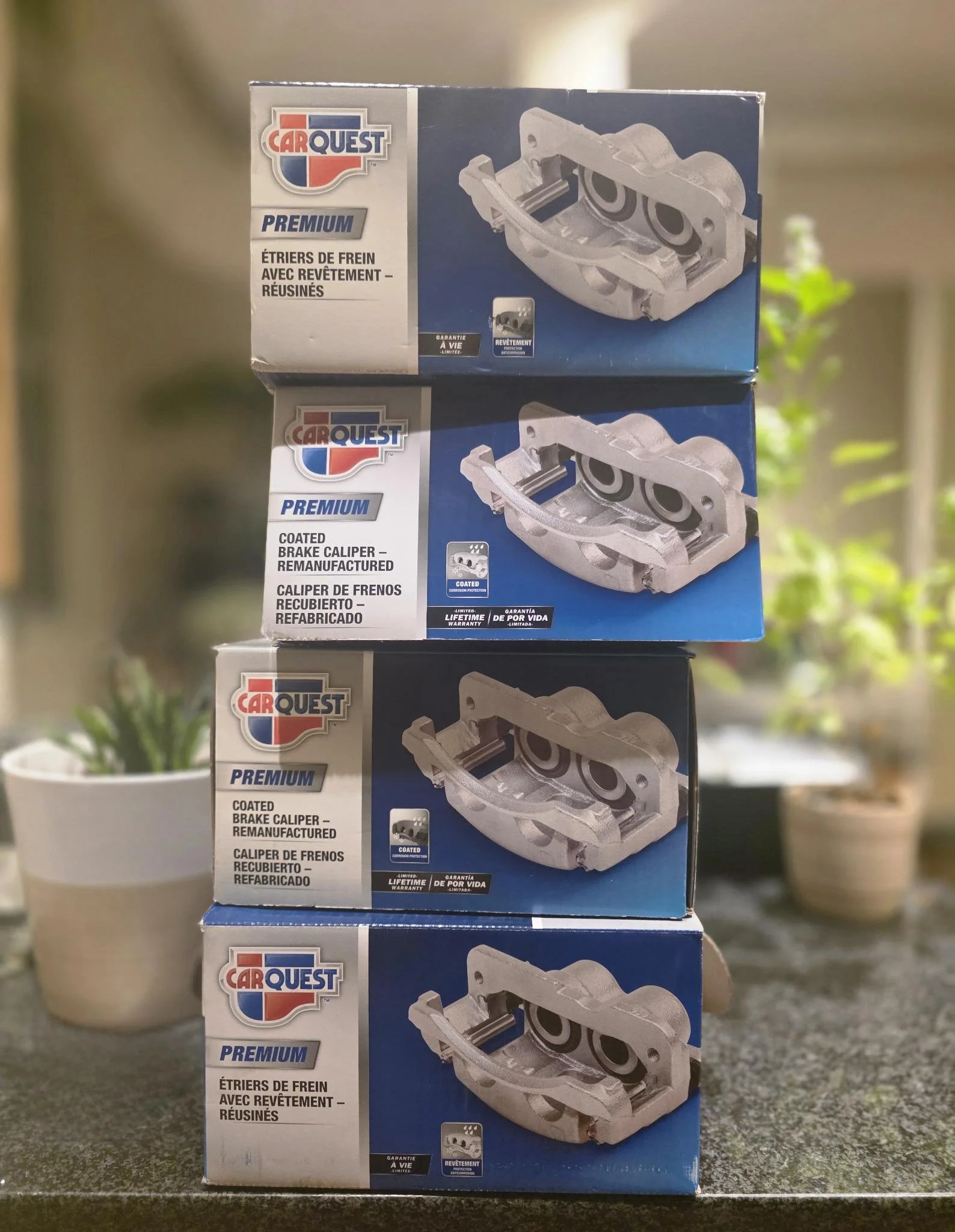 Four boxes of Carquest premium coated brake calipers stacked on a countertop with potted plants in the background.