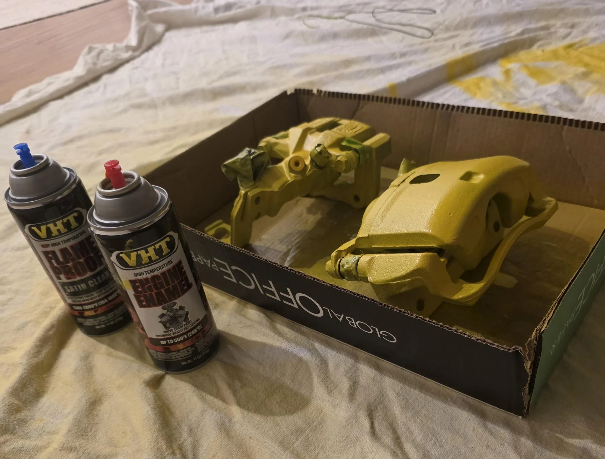 Yellow painted brake calipers inside a cardboard box on a table with spray paint cans nearby.