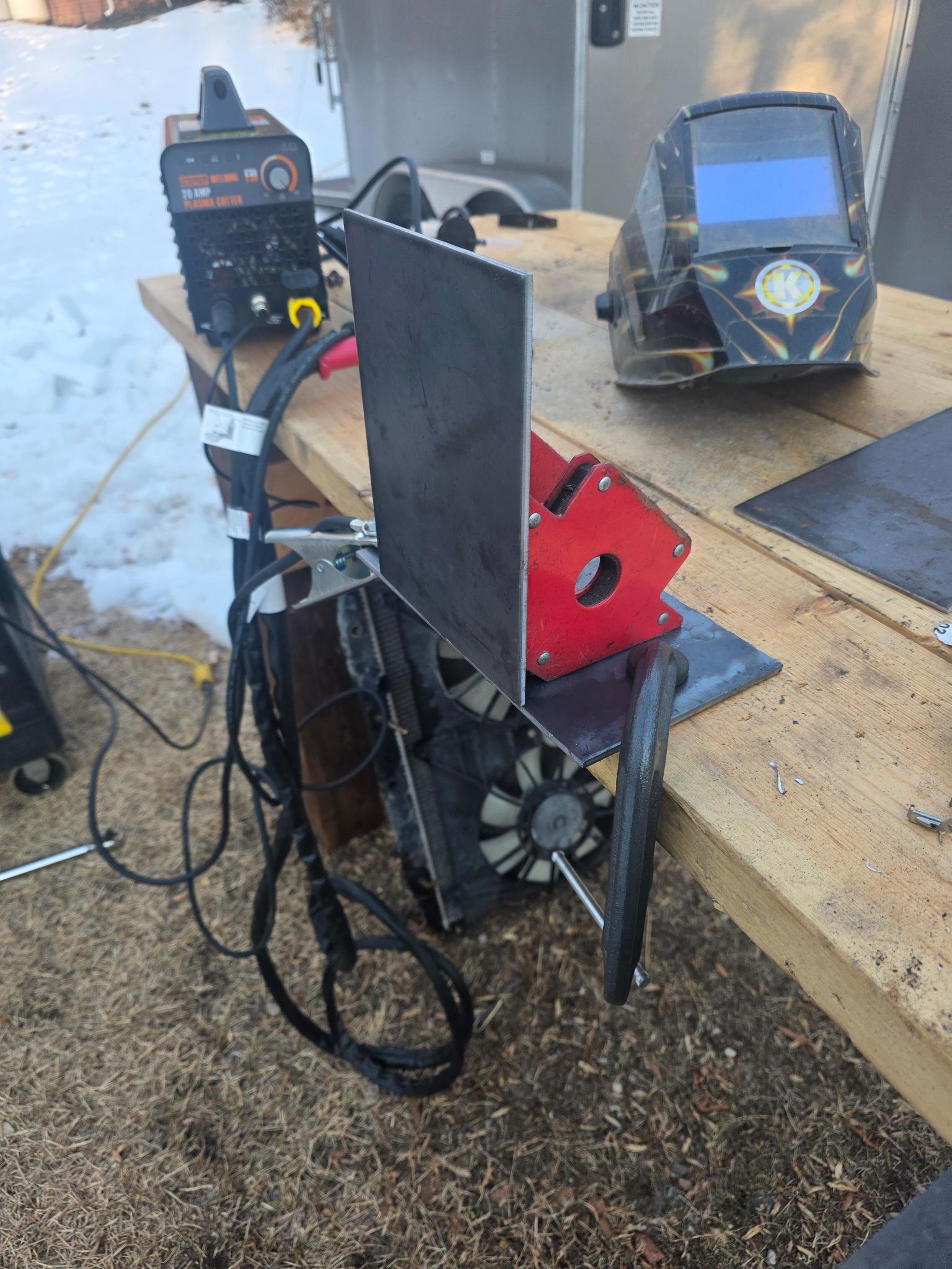 A welding workspace outdoors with a welding helmet, welding machine, and a metal workpiece clamped to a wooden work surface.