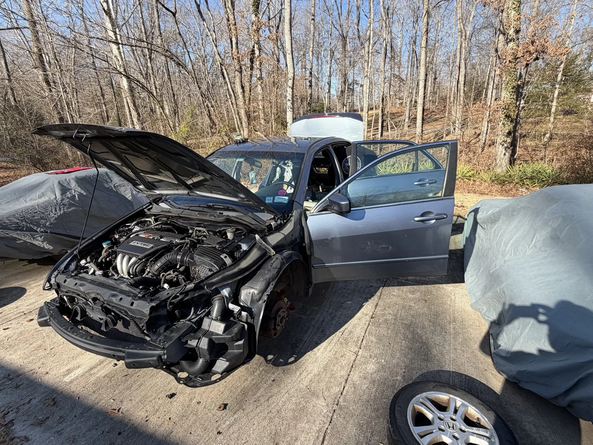 A gray car with an open hood and the front wheel removed, parked outdoors on a concrete surface with other covered vehicles around, in a wooded area during daylight.