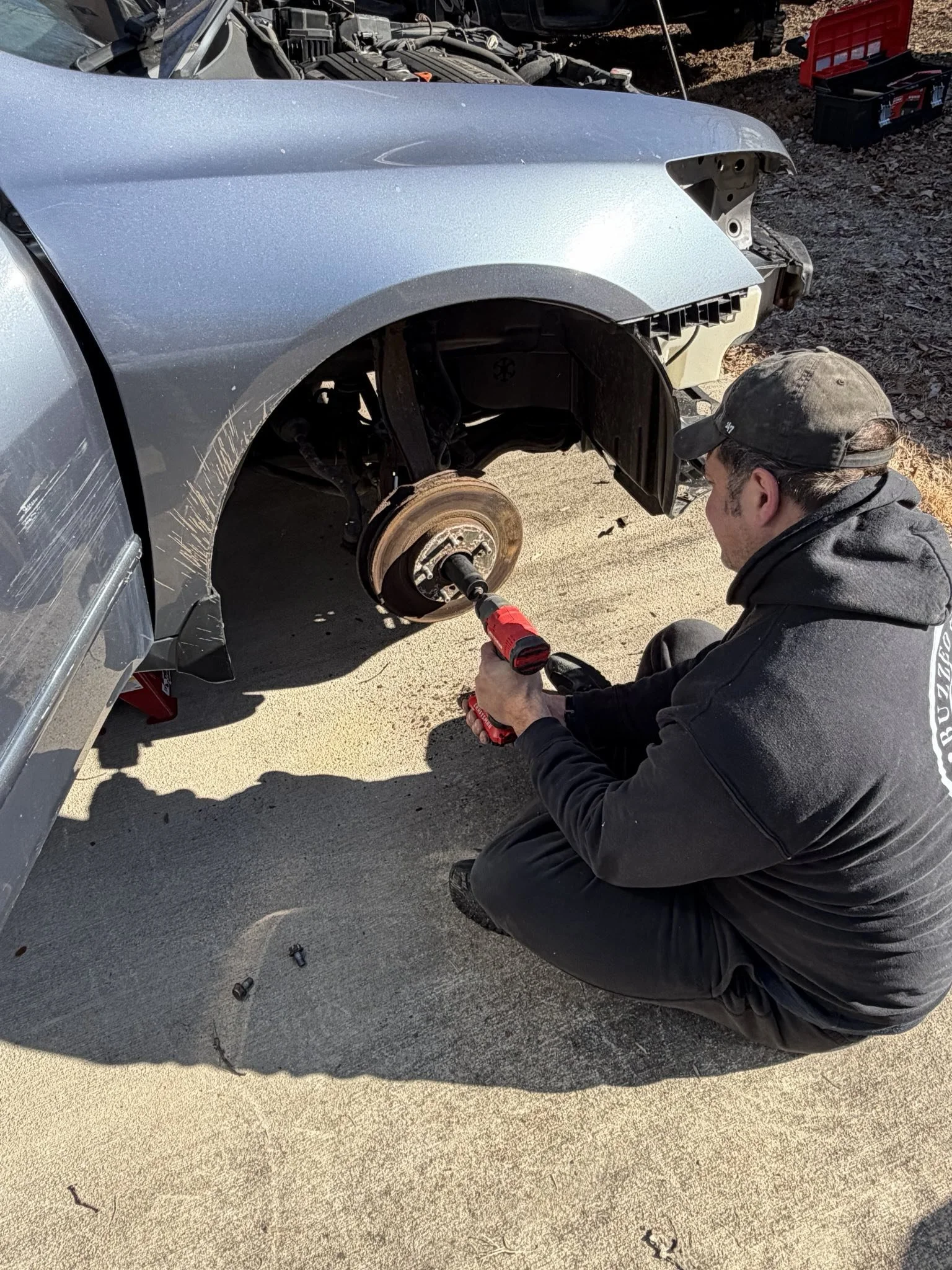 Person working on a car's front wheel assembly, using a cordless drill, with the wheel removed and brake components visible.