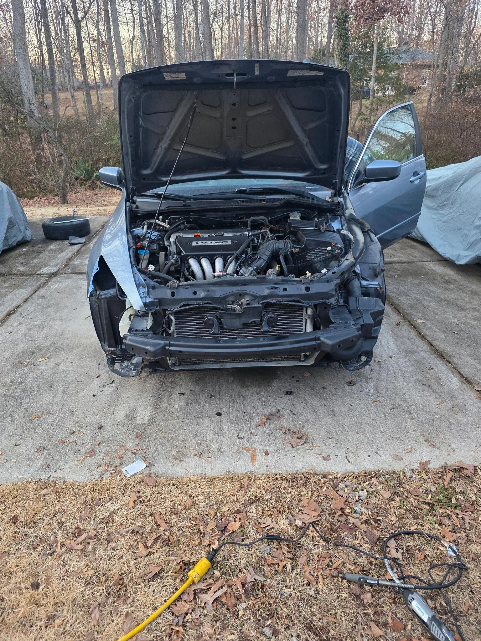 A car with its hood open and front bumper removed, exposing the engine and radiator, parked on a concrete driveway with tools and parts nearby.