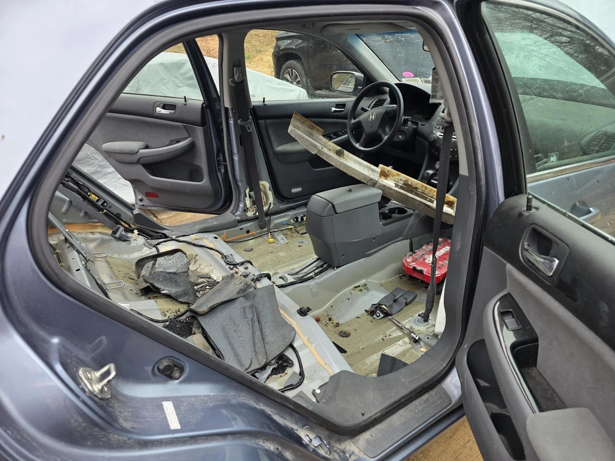 The interior of a  2007 Honda Accord with no seats, showing exposed floor and dashboard, with various debris and tools inside in prepapration for roll cage installation.