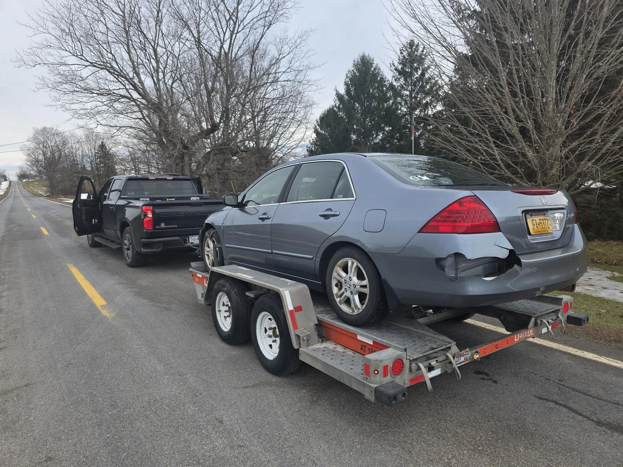 A gray sedan with rear bumper damage on a U-Haul trailer attached to a black pickup truck, parked on the side of a rural road with leafless trees and cloudy sky in the background.