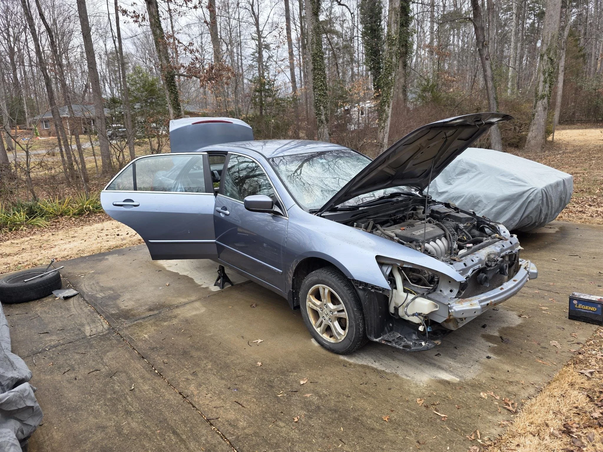 A blue sedan with its hood open, missing front bumper, and one front wheel removed, parked on a driveway surrounded by trees, with a covered vehicle behind it.