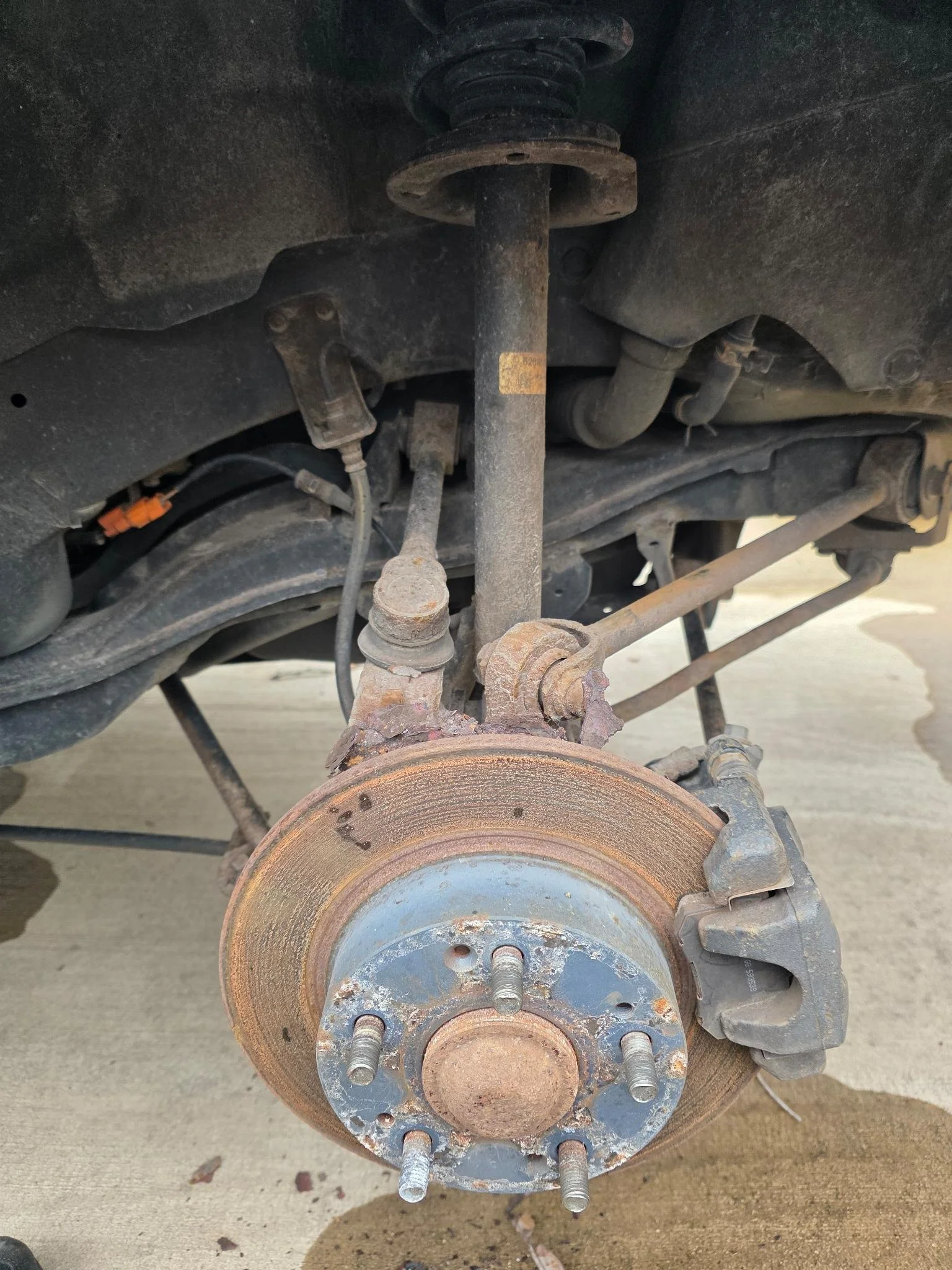 Car's front wheel removed, showing rusty brake rotor, brake caliper, and suspension components.