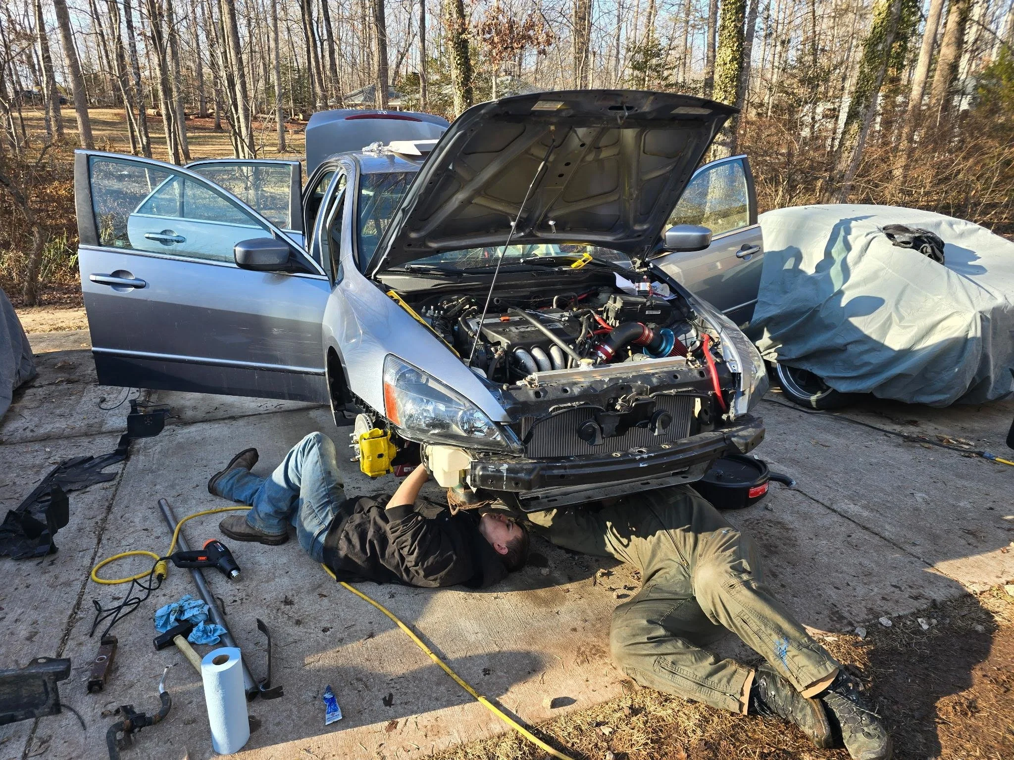 Two mechanics working underneath the front of a silver car with its hood open on a driveway surrounded by trees. There are tools and equipment scattered nearby, and another car is covered with a tarp to the right.