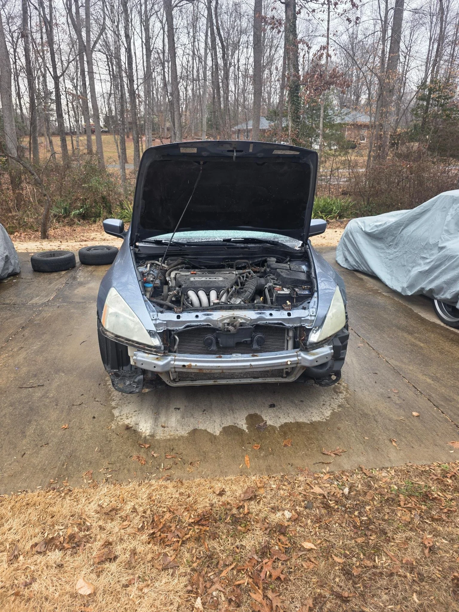 Front view of a silver car with its hood open, showing the engine and internal components, parked on a driveway with a wet patch underneath, with covered vehicles and leafless trees in the background.