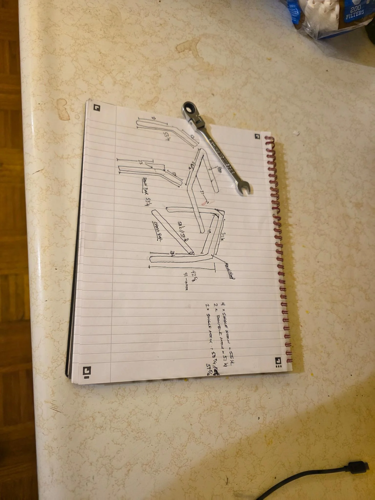 A sketch with measurements and annotations on lined notebook paper, accompanied by an open-end wrench placed on the paper. The paper is on a beige, textured surface with some stains.