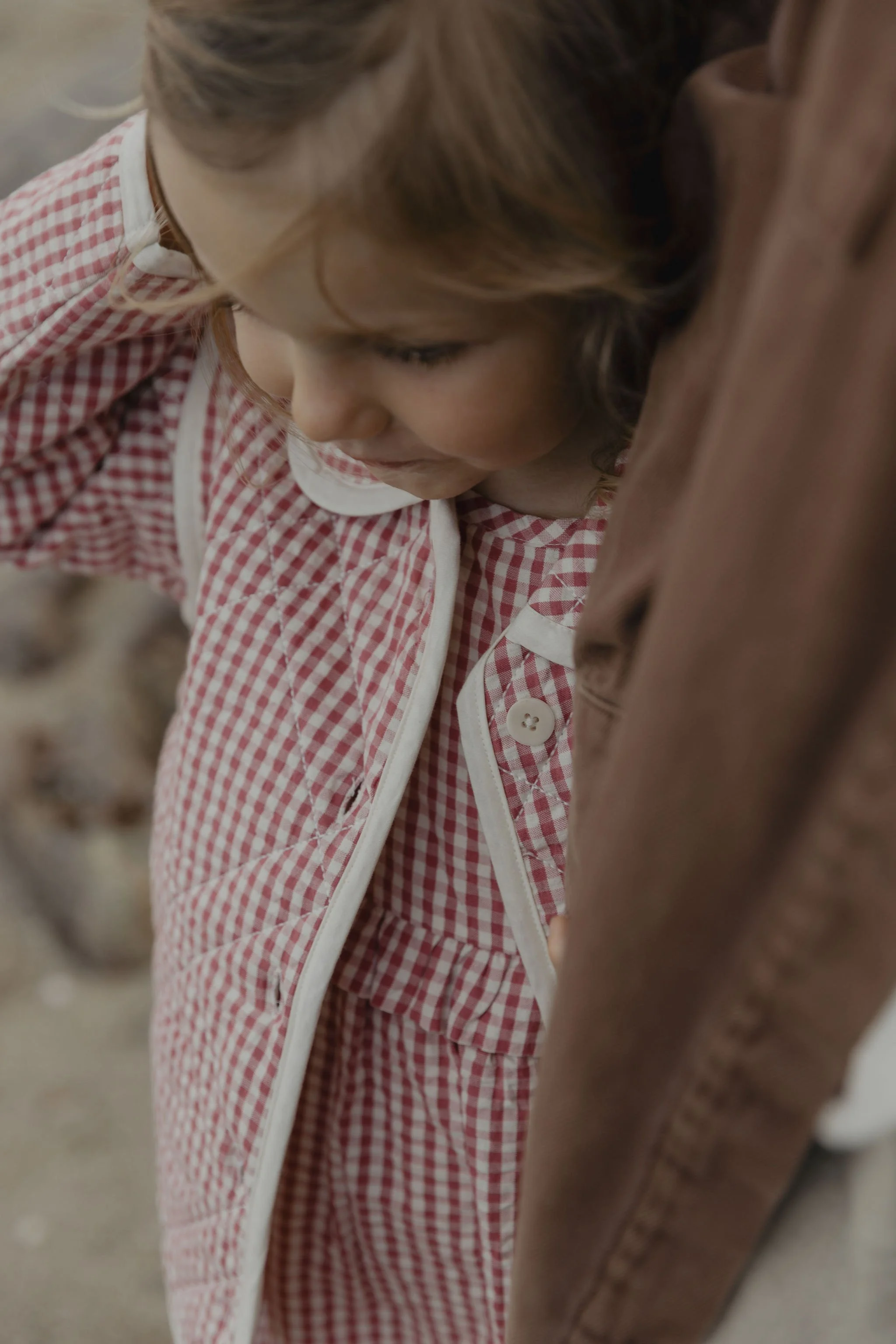 Child in red and white checkered dress with white trim looking downward, leaning against a brown leather surface.